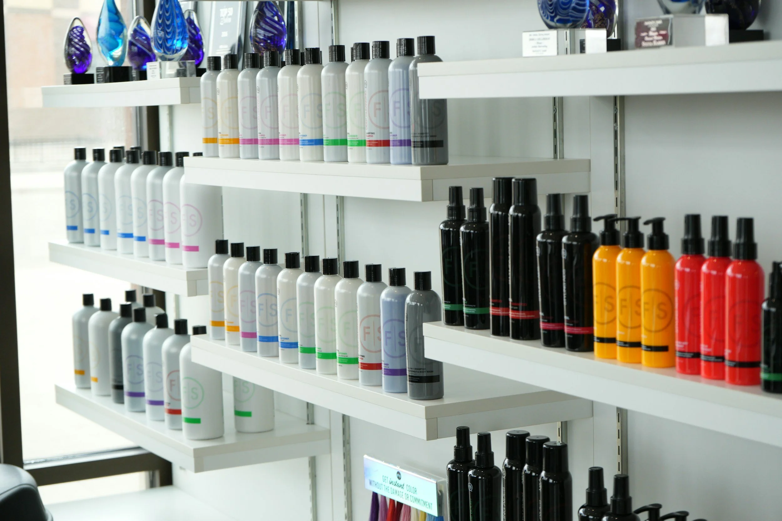 White shelves holding various bottles of colorful hair care products, including shampoos and sprays, in a retail store.