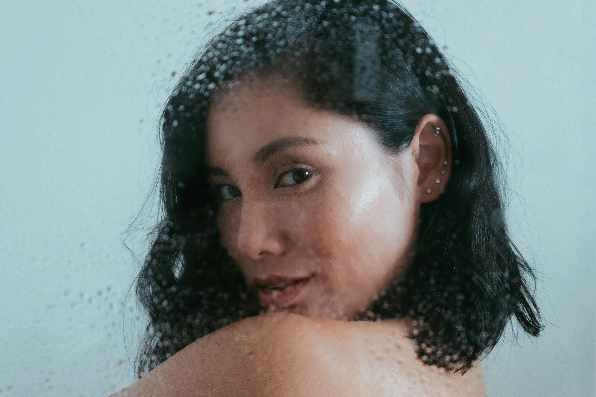Close-up of a woman with black hair, brown eyes, and multiple piercings in her ear, looking into a mirror with water droplets on it.