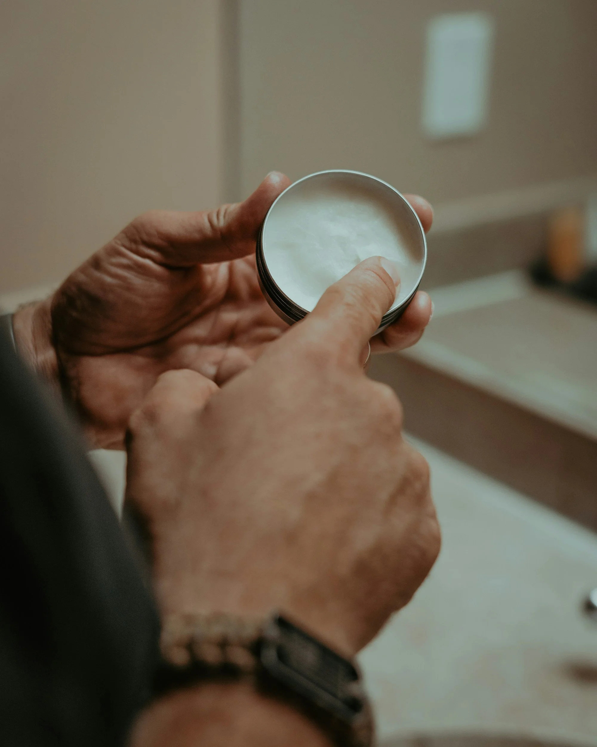 A hand holding a round metal container with a white substance inside, possibly lotion or cream.