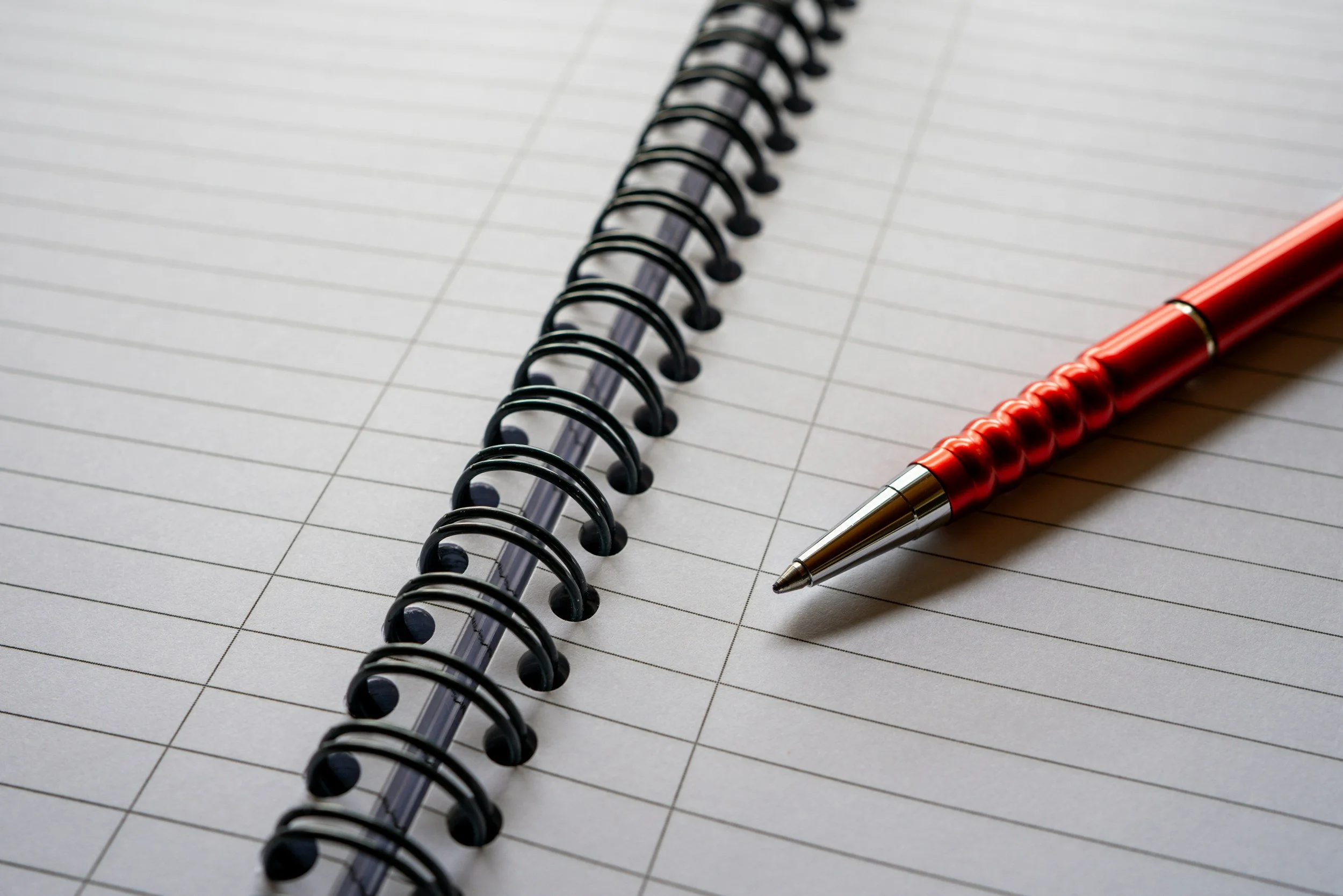 A red and silver pen resting on an open blank spiral notebook with lined pages.