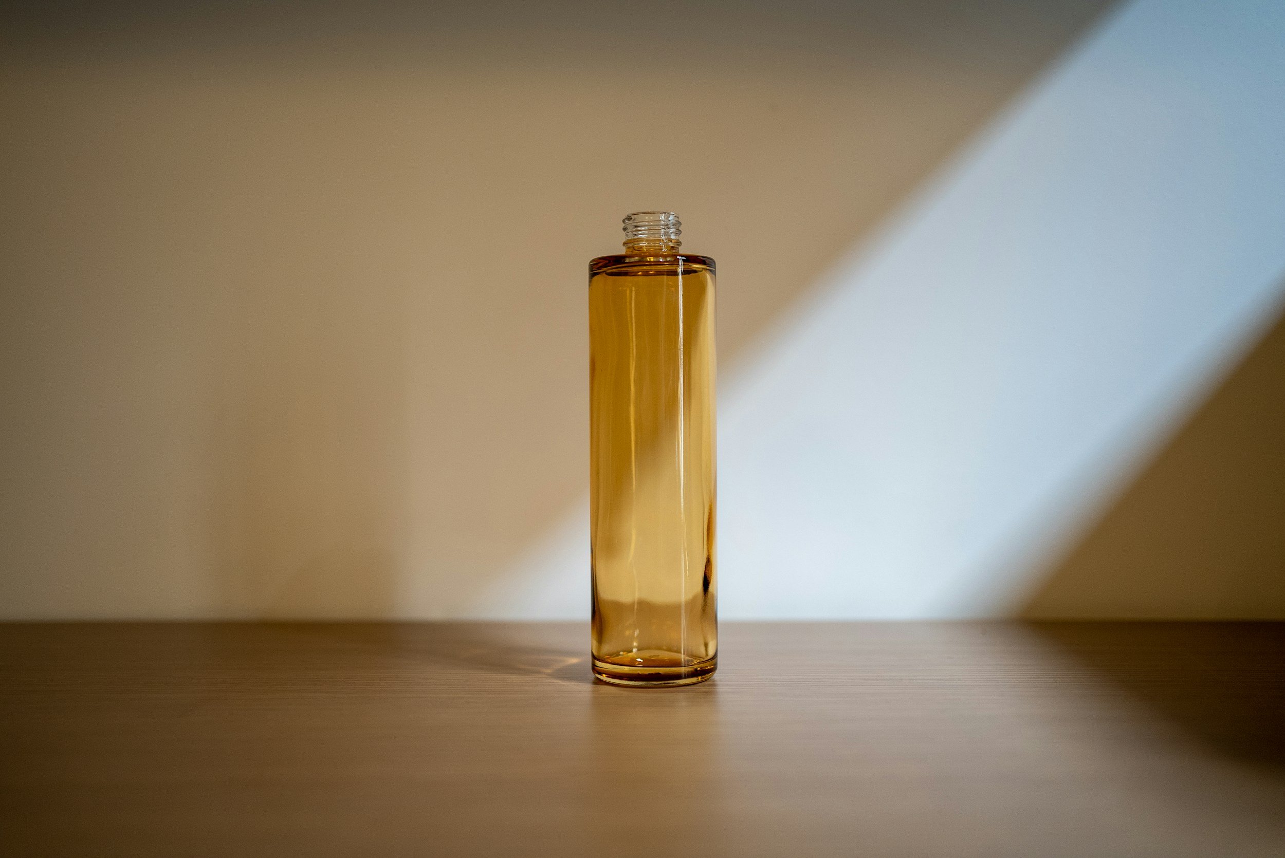 A tall, transparent glass bottle filled with amber-colored liquid, placed on a flat surface with a sharp shadow cast behind it.