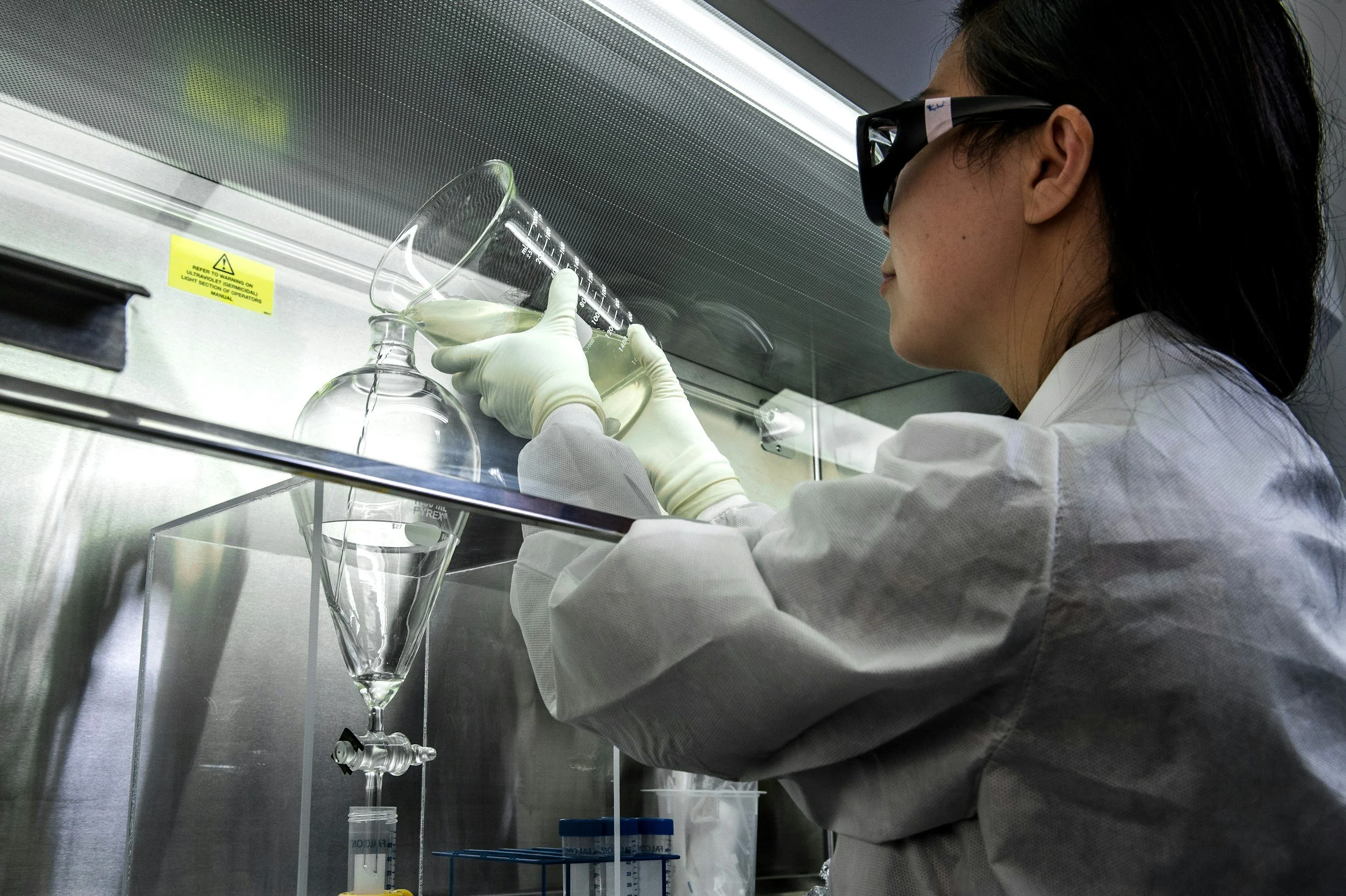 A scientist wearing safety goggles and gloves pouring a liquid into a laboratory glassware inside a fume hood.