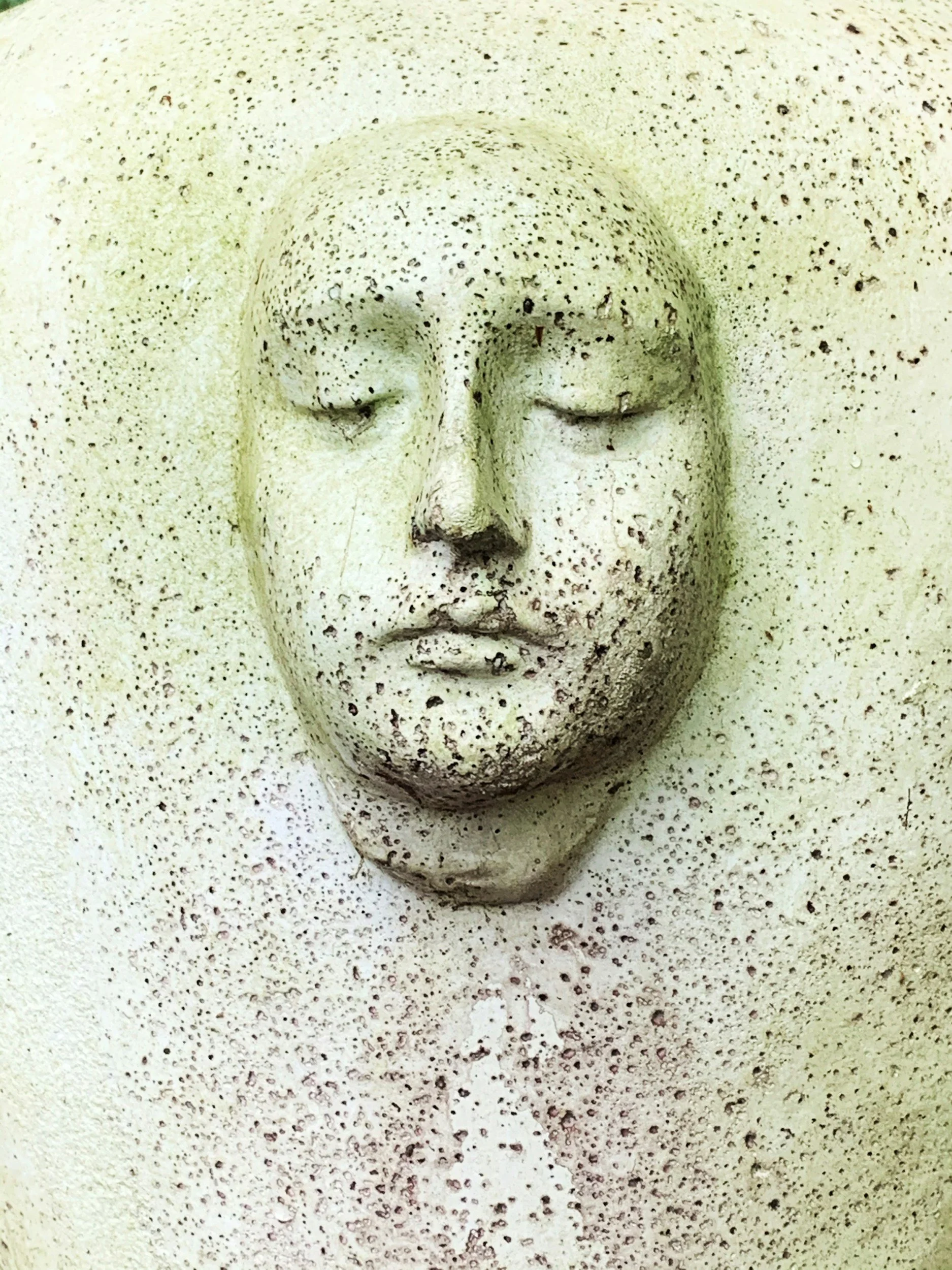Close-up of a speckled concrete or stone surface with a carved face sculpture showing closed eyes and lips.