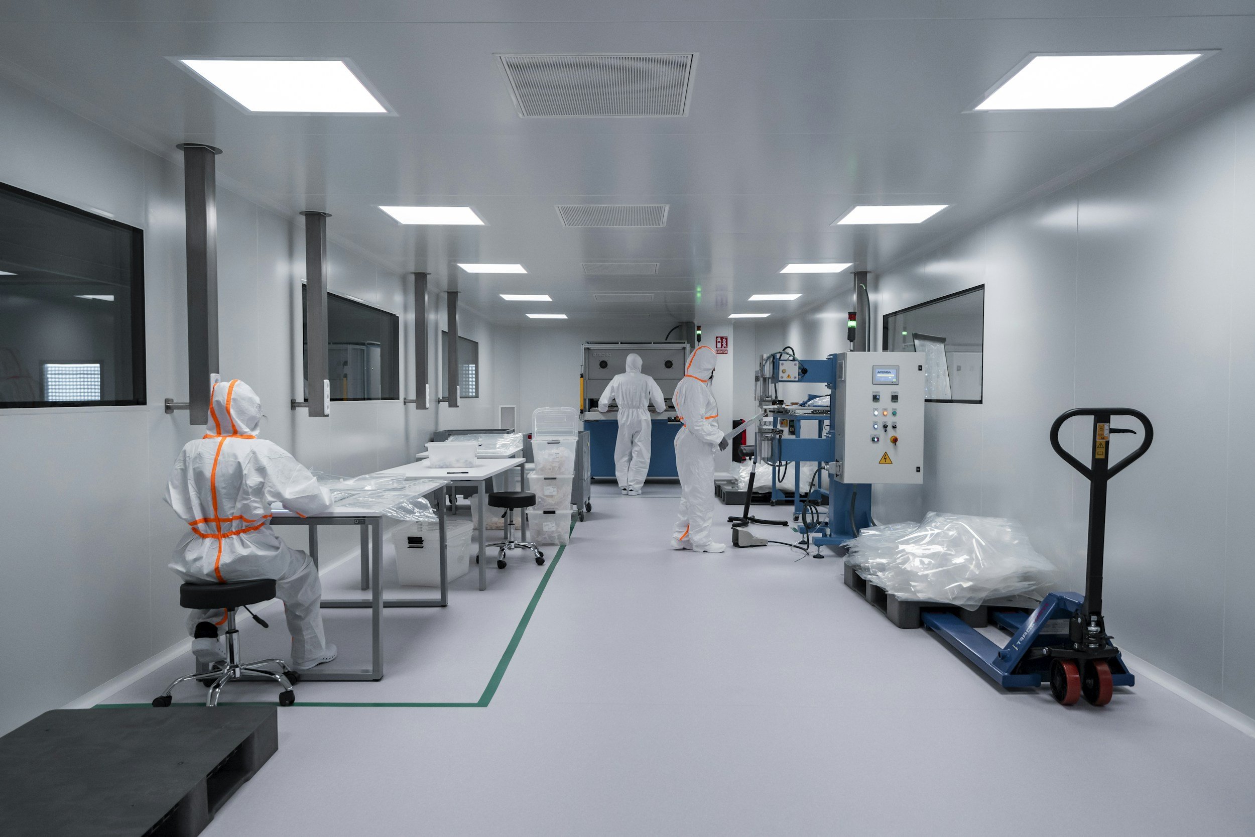 A sterile cleanroom laboratory with four workers dressed in white protective suits with orange stripes. They are working with scientific or manufacturing equipment, including electronic machines and laboratory supplies in a modern, well-lit environment.