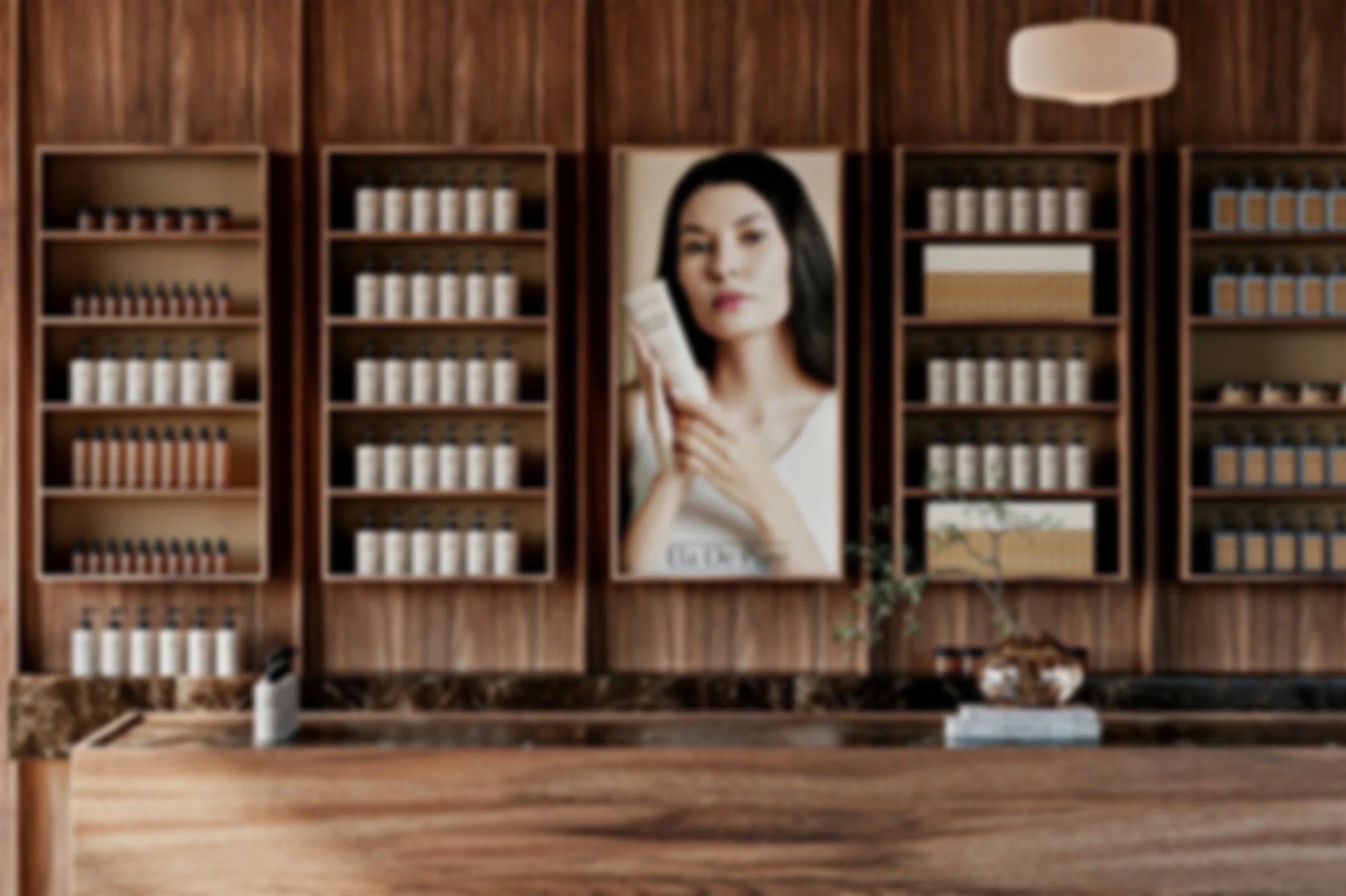 A woman with shoulder-length black hair holds a small container and gazes thoughtfully. She is positioned in front of a wooden shelving unit with multiple rows of jars and boxes, and a white wall-mounted lamp is above the shelves.