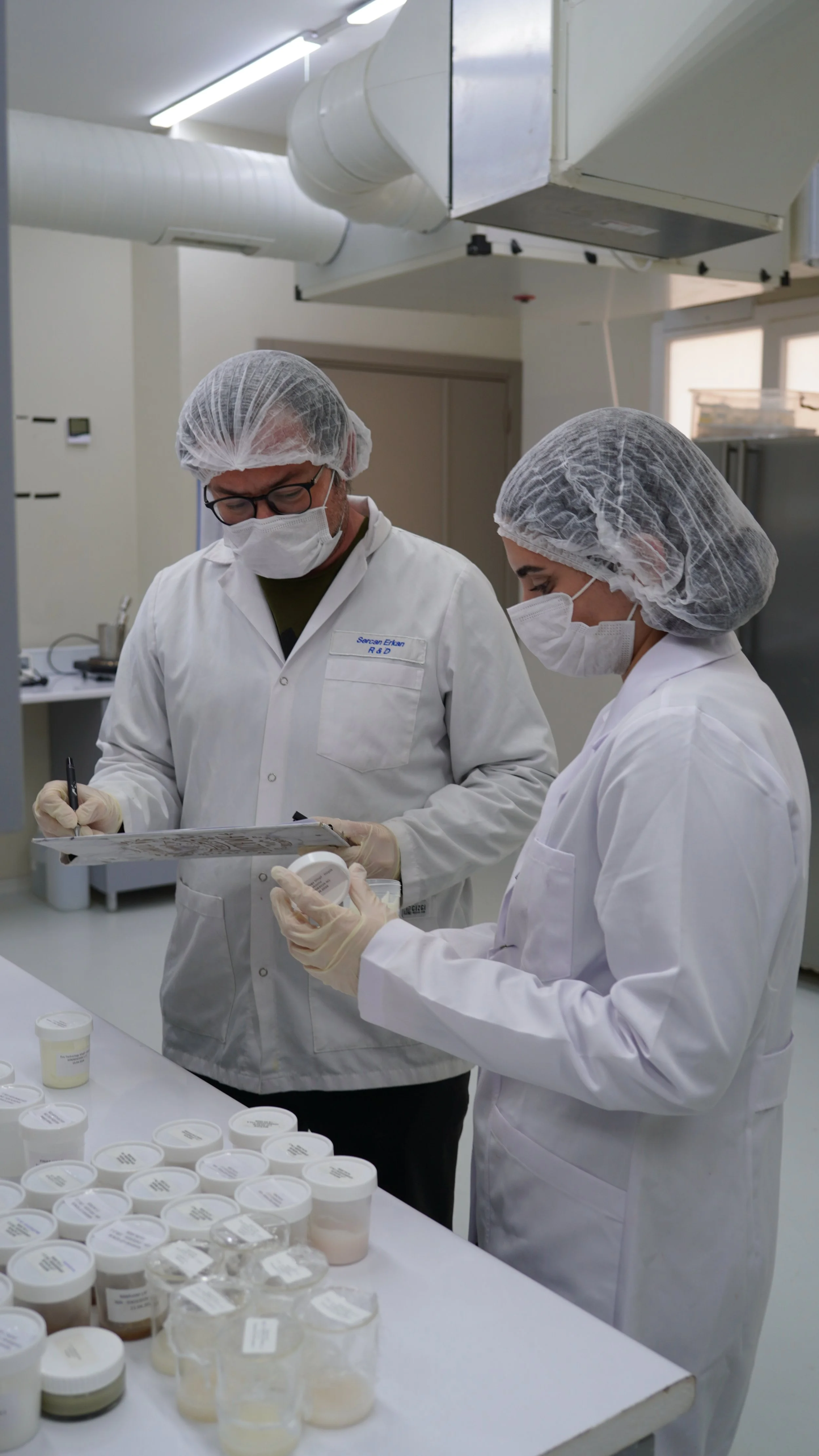 Two scientists wearing lab coats, masks, hairnets, and gloves examining samples in a laboratory surrounded by containers with labels.