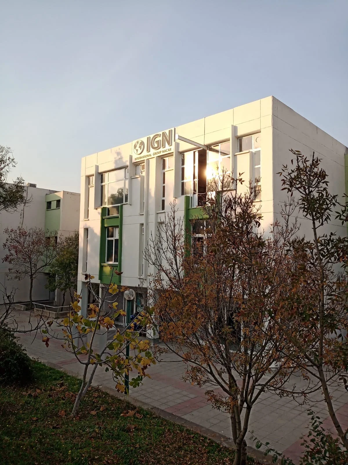 Photo of a modern white building with green accents, labeled 'IGN International Group NACAR' at the top, with trees and a sidewalk in the foreground, during sunset or late afternoon.