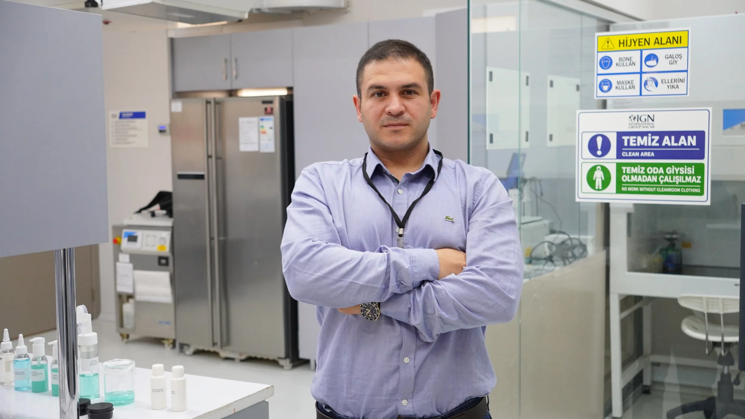 Gri saçlı, mavi kazak ve bej pantolon giymiş bir adam, ellerini bankın üzerine koymuş halde oturuyor ve sade bir arka planın önünde yan tarafa bakıyor.