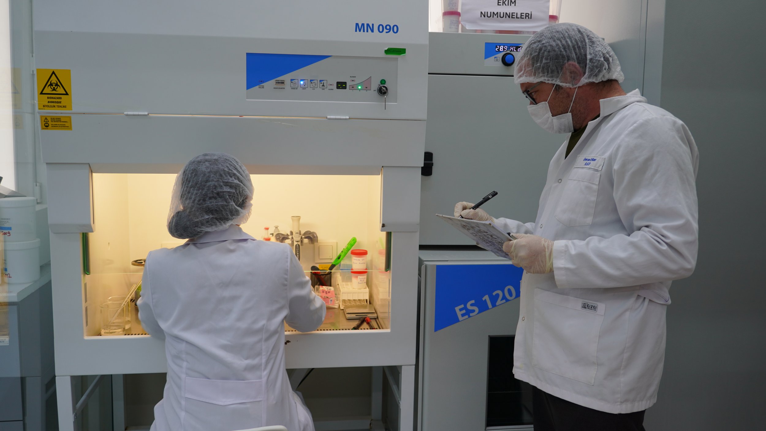 Scientists working in a laboratory with a biosafety cabinet. One scientist is inside the cabinet working with lab equipment, while another scientist is standing outside, taking notes or observing. Both are wearing protective clothing, masks, and hairnets.