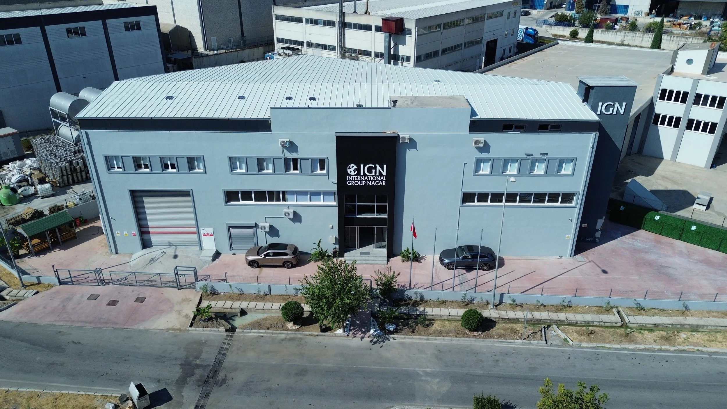 An aerial view of a modern industrial building with a gray exterior, multiple windows, and a large garage door. The building has the signs "IGN" and "International Group NACAR" on the front. There are two parked cars in front and a paved sidewalk with nearby trees and equipment around the building.