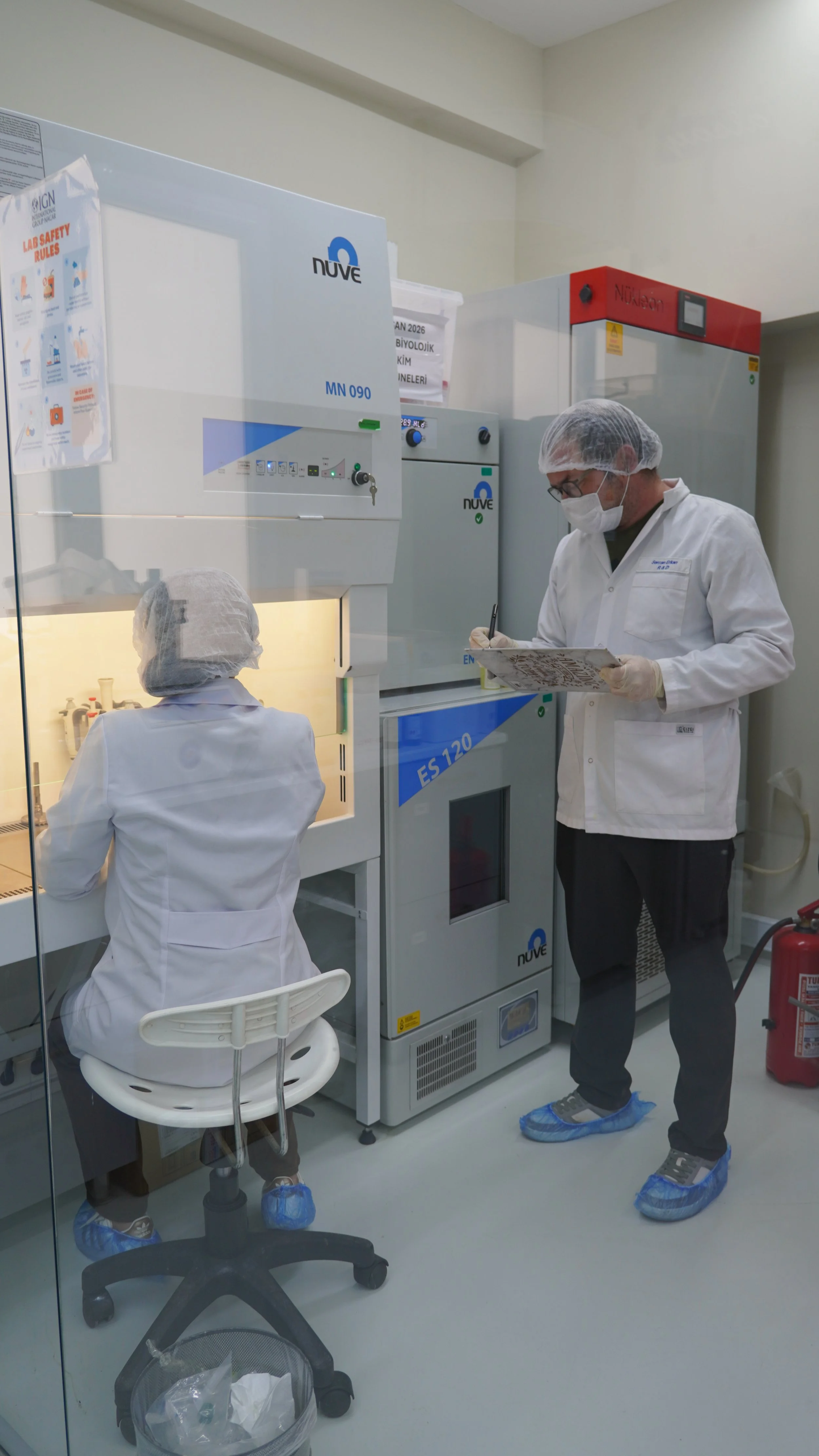 Two scientists in protective clothing working in a laboratory. One scientist is sitting in front of a biological safety cabinet, and another is standing with a clipboard, both wearing gloves, masks, hair covers, and shoe covers.