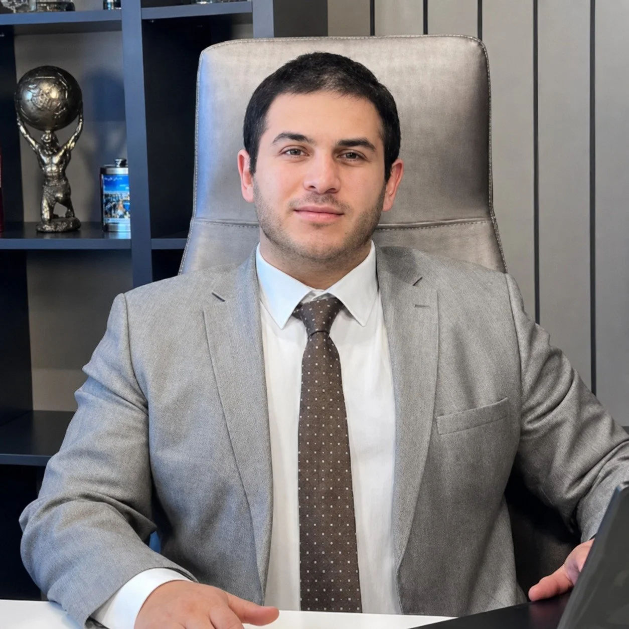 Gri saçlı, mavi kazak ve bej pantolon giymiş bir adam, ellerini bankın üzerine koymuş halde oturuyor ve sade bir arka planın önünde yan tarafa bakıyor.