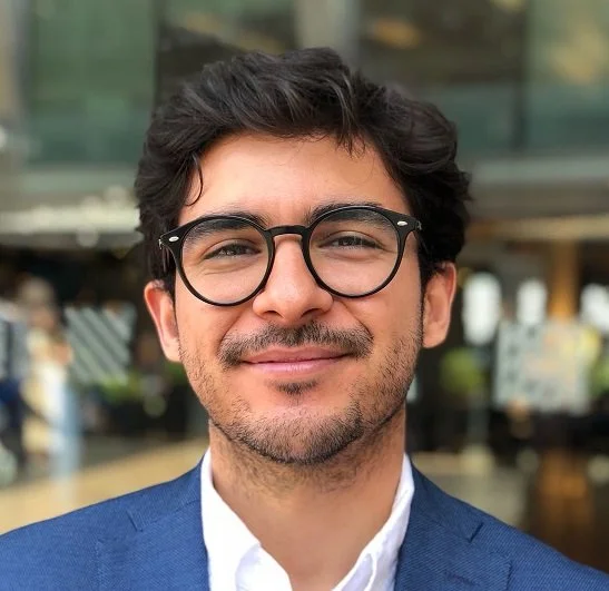 Young man with glasses smiling in an urban setting.