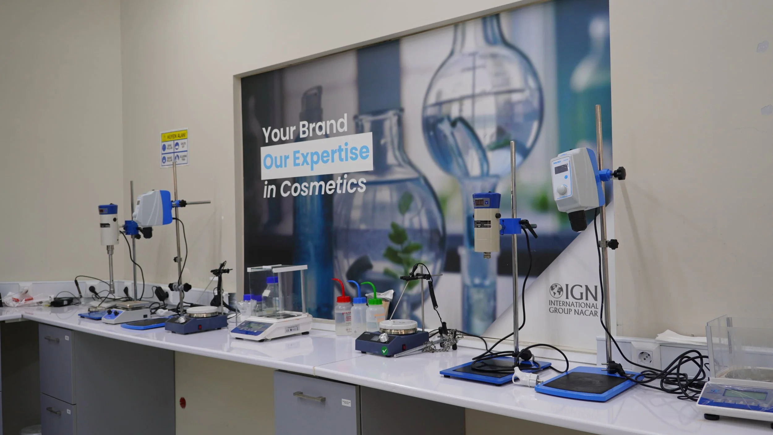 A science laboratory setup with various scientific instruments and equipment on a white counter, including scales, pipettes, and glass containers, with a large banner in the background displaying scientific images and the text "Your Brand Our Expertise in Cosmetics."