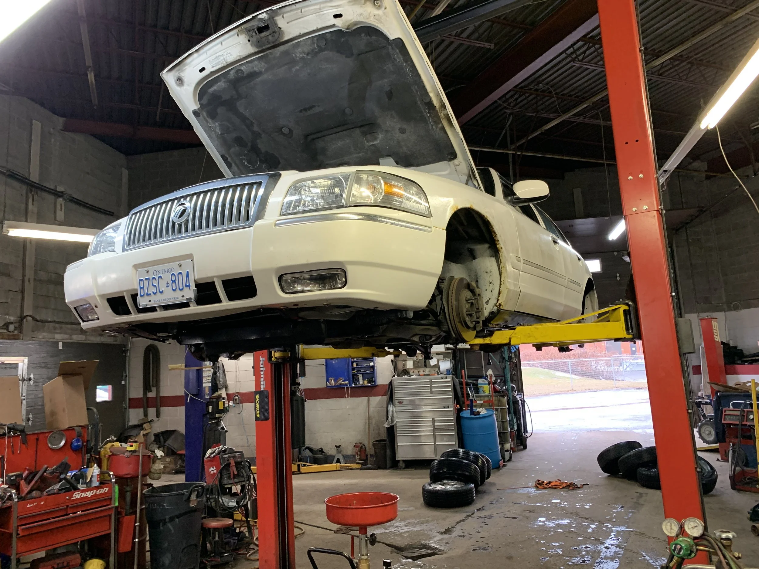 A white Cadillac vehicle on a hydraulic lift inside an auto repair shop, with its hood open, tools, and tires scattered around.