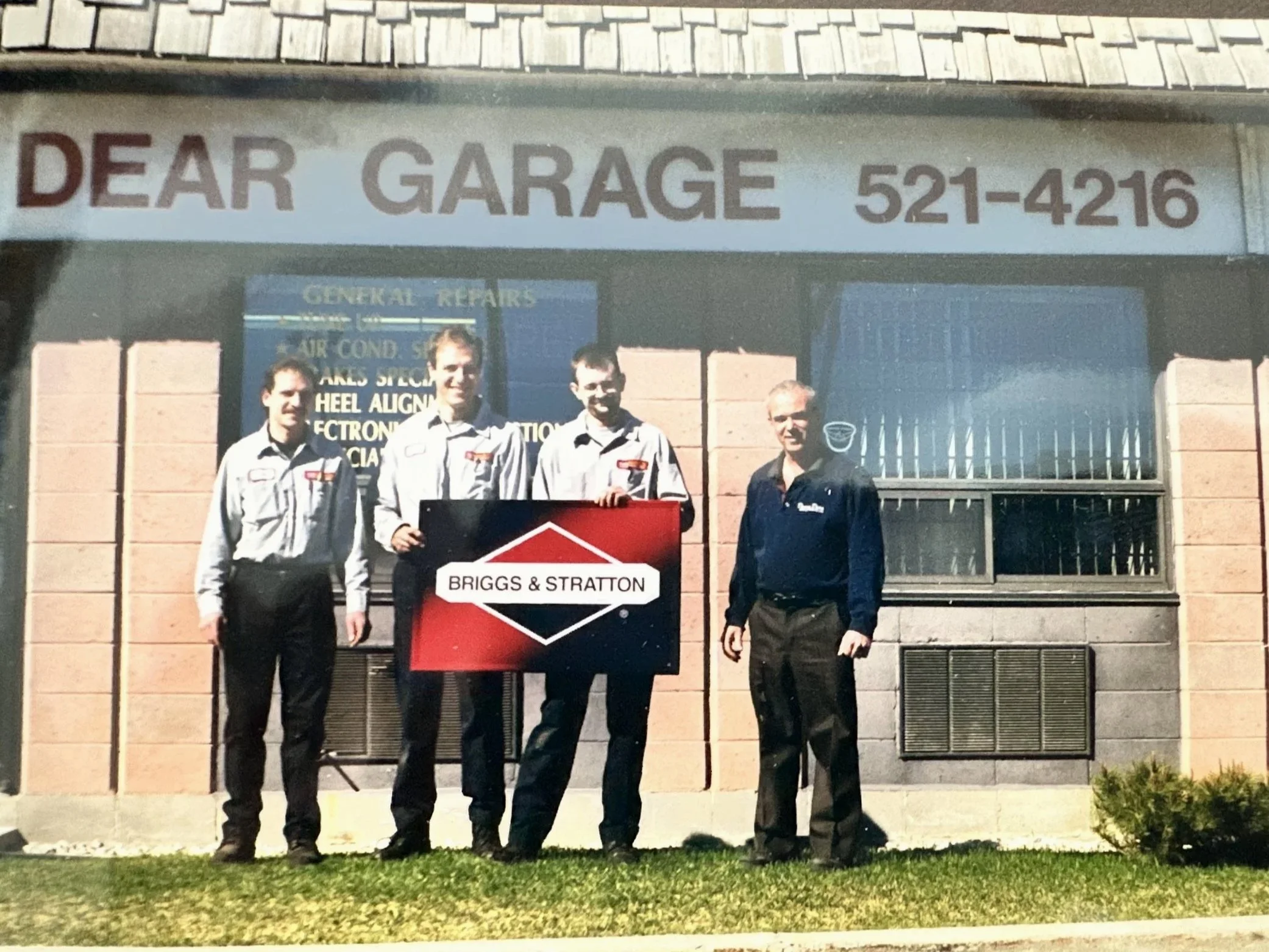Four men standing outside a building with a sign that reads "Dear Garage" and a phone number. One man is holding a sign that says "Briggs & Stratton."