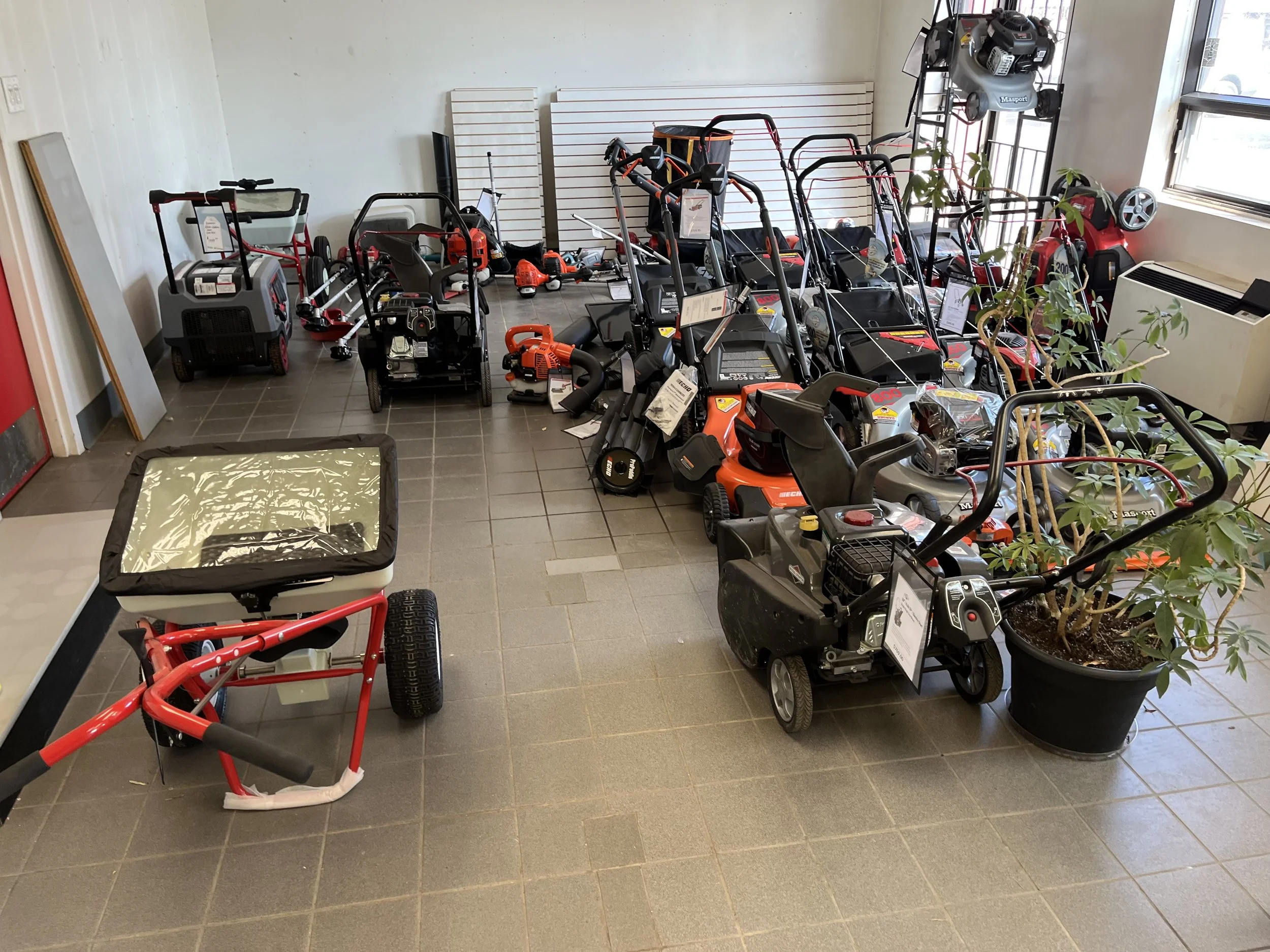Display of various outdoor power equipment including lawnmowers, trimmers, and blowers, along with some gardening tools, arranged indoors near windows with a white slatted wall panel in the background.