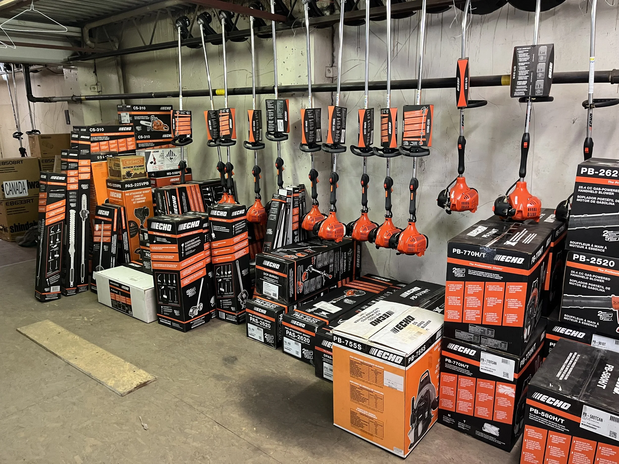 A display of power tools, mainly leaf blowers, in orange and black, on a store shelf. Various tool boxes and packaging are stacked below and around the tools.