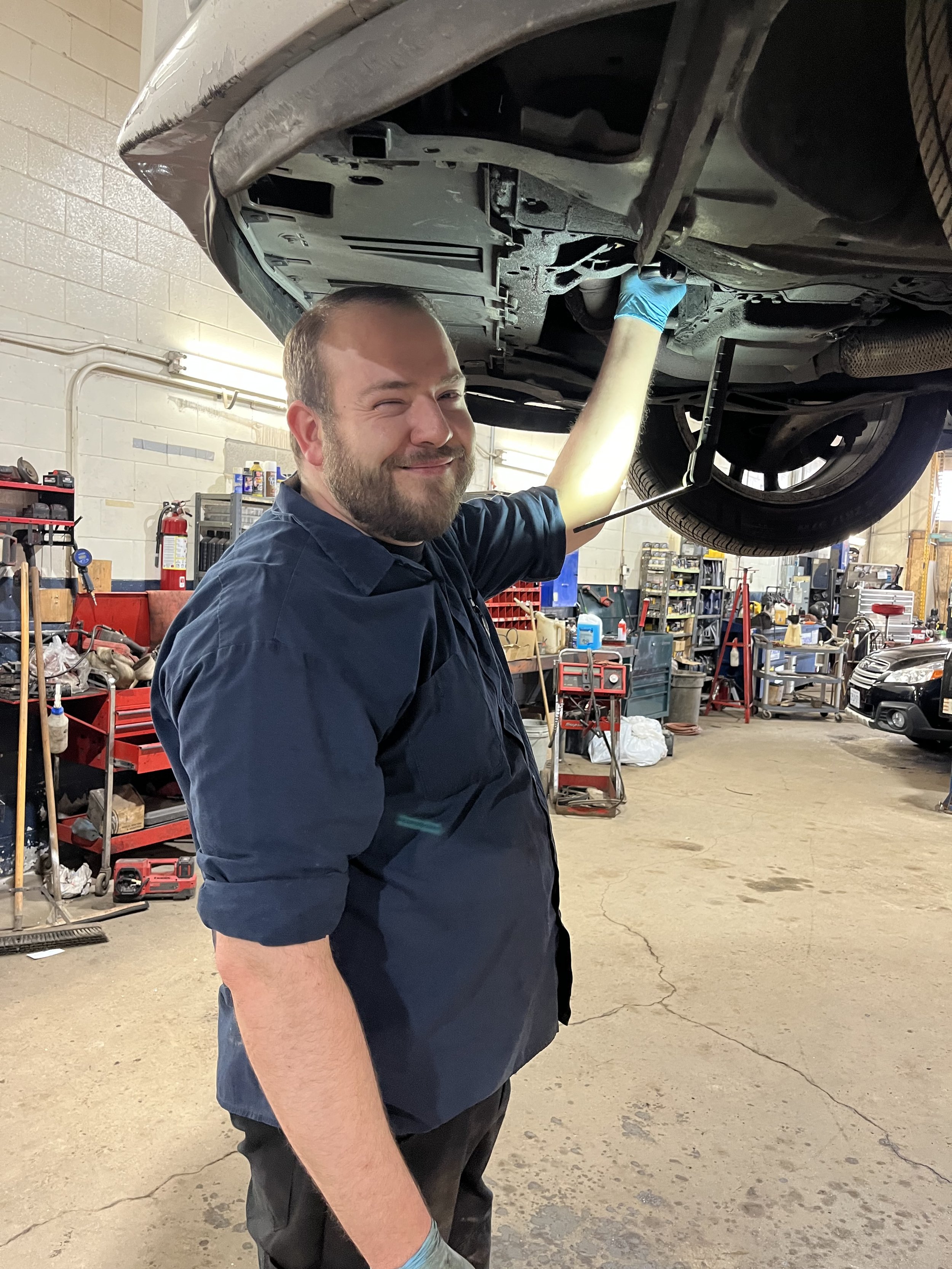 A man wearing a dark blue mechanic uniform and gloves is working underneath a lifted vehicle in an auto repair shop. The shop has shelves with tools and supplies and other vehicles are visible in the background.