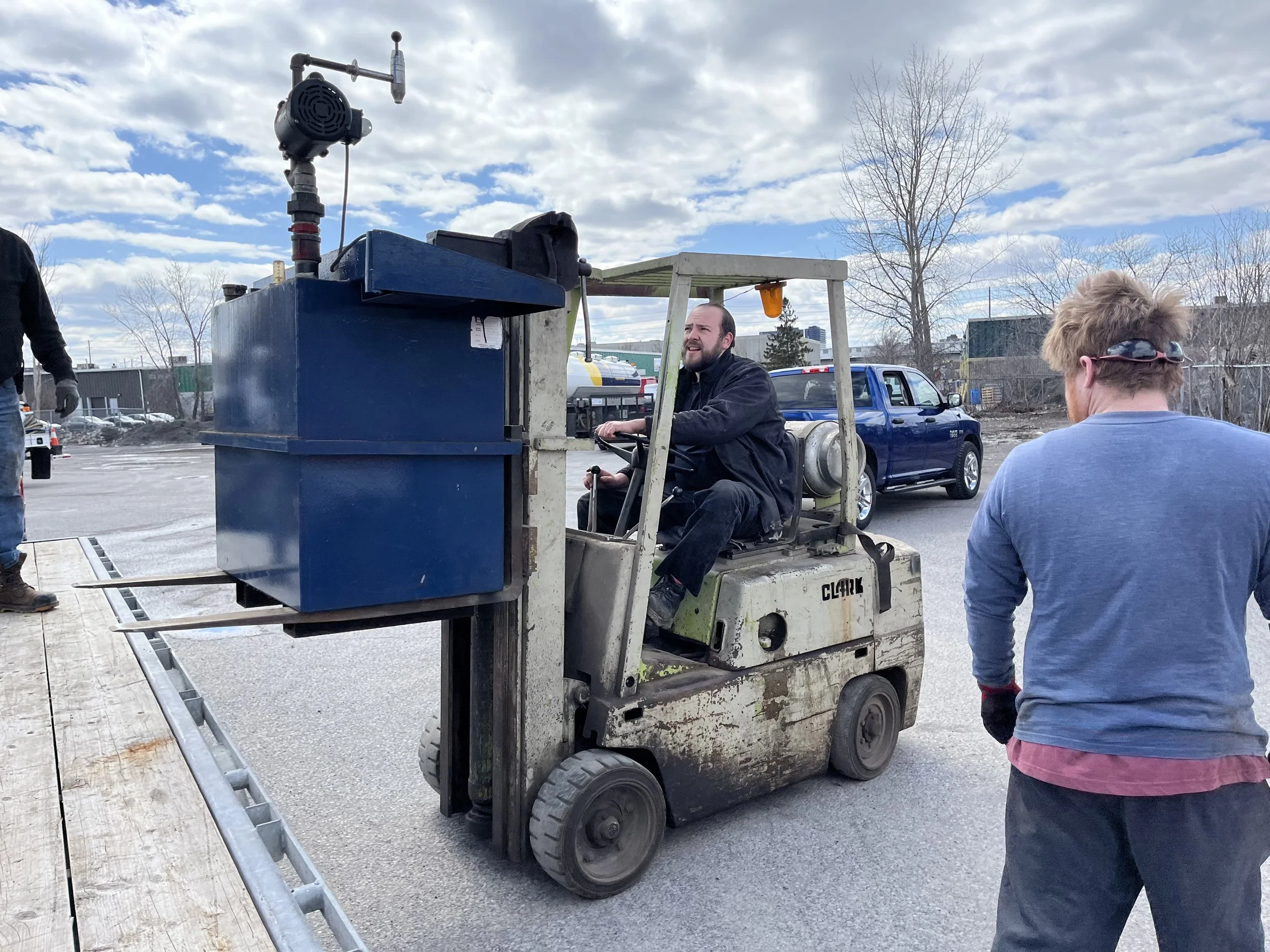 A man operating a forklift machine outdoors during daytime, with a woman with sunglasses nearby, a car, and leafless trees in the background under a partly cloudy sky.