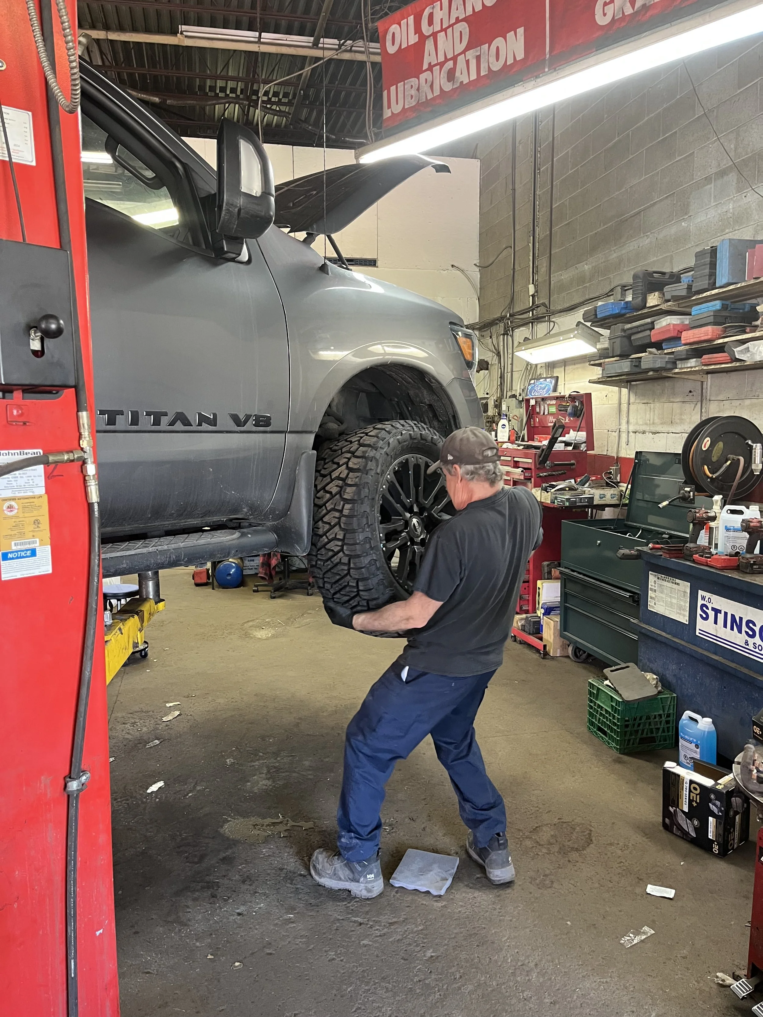 A mechanic working on a lifted gray Toyota Titan V8 truck inside an auto repair shop. The truck is on a hydraulic lift, and the mechanic is maneuvering a large tire or wheel. The shop has various tools, an organized shelf with supplies, and a sign overhead that reads 'OIL CHANGE AND LUBRICATION'.