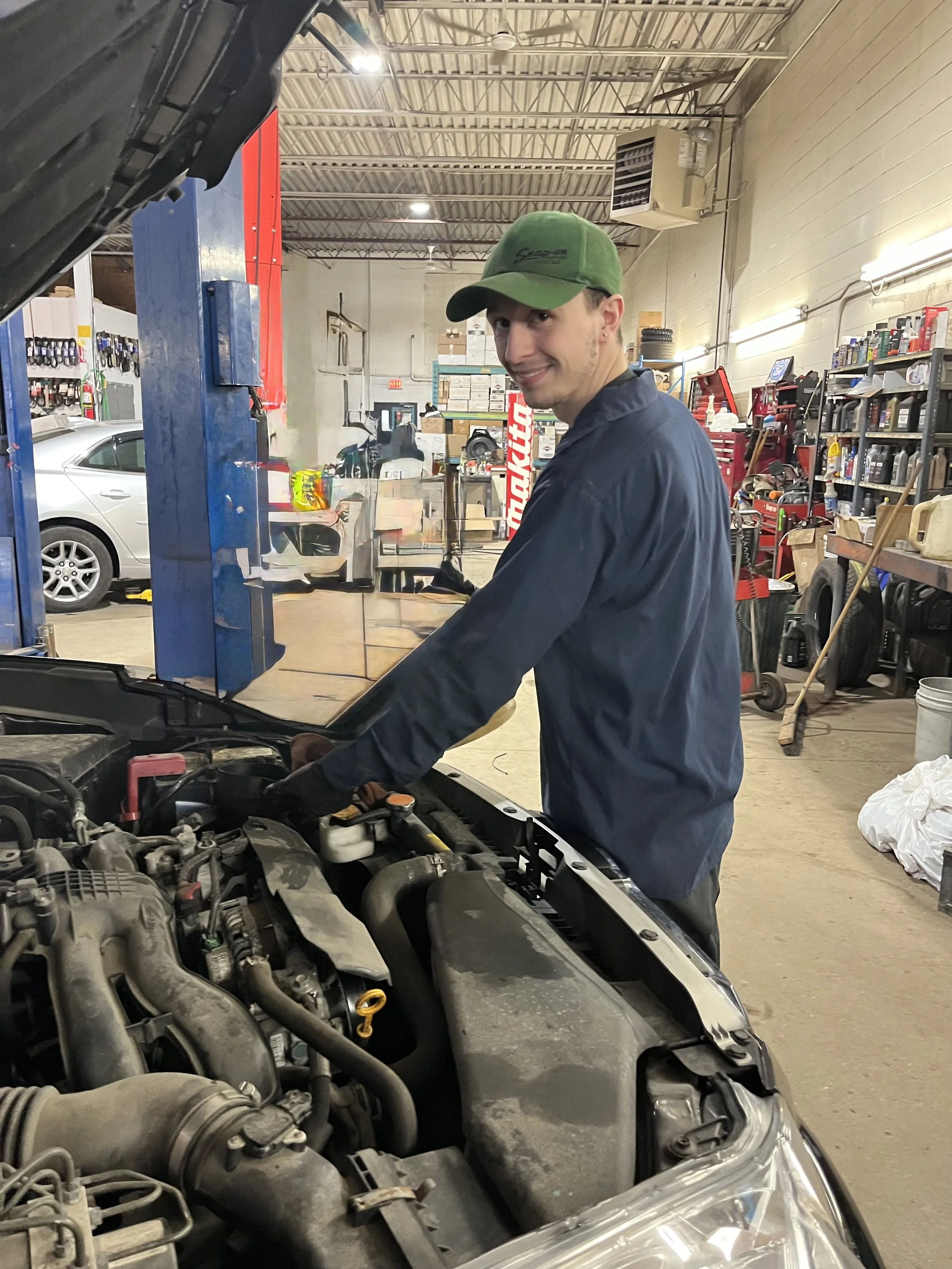 A young man wearing a green cap and dark jacket working on a car in an auto repair shop, smiling at the camera, with various tools and shelves in the background.