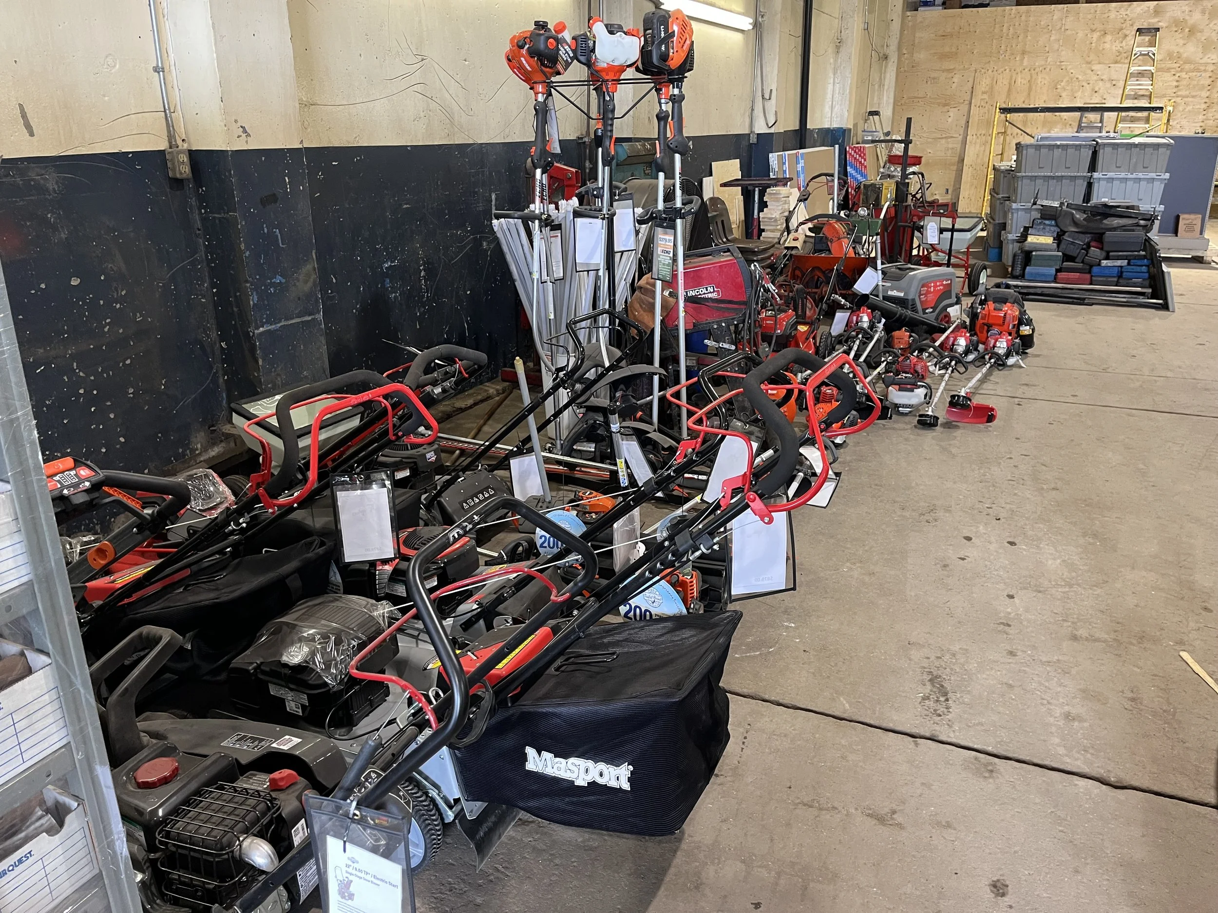 Various outdoor power tools and equipment, including lawnmowers, trimmers, and leaf blowers, are displayed for sale in a hardware store aisle.