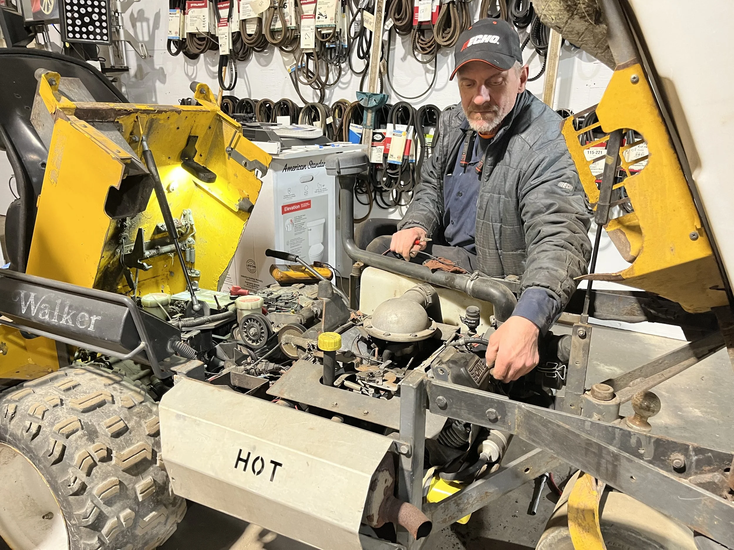 A man wearing a black cap and gray jacket working on a yellow construction vehicle in a hardware store. The store has belts hanging on the wall in the background. The vehicle has a sign that reads 'HOT' on the side panel.
