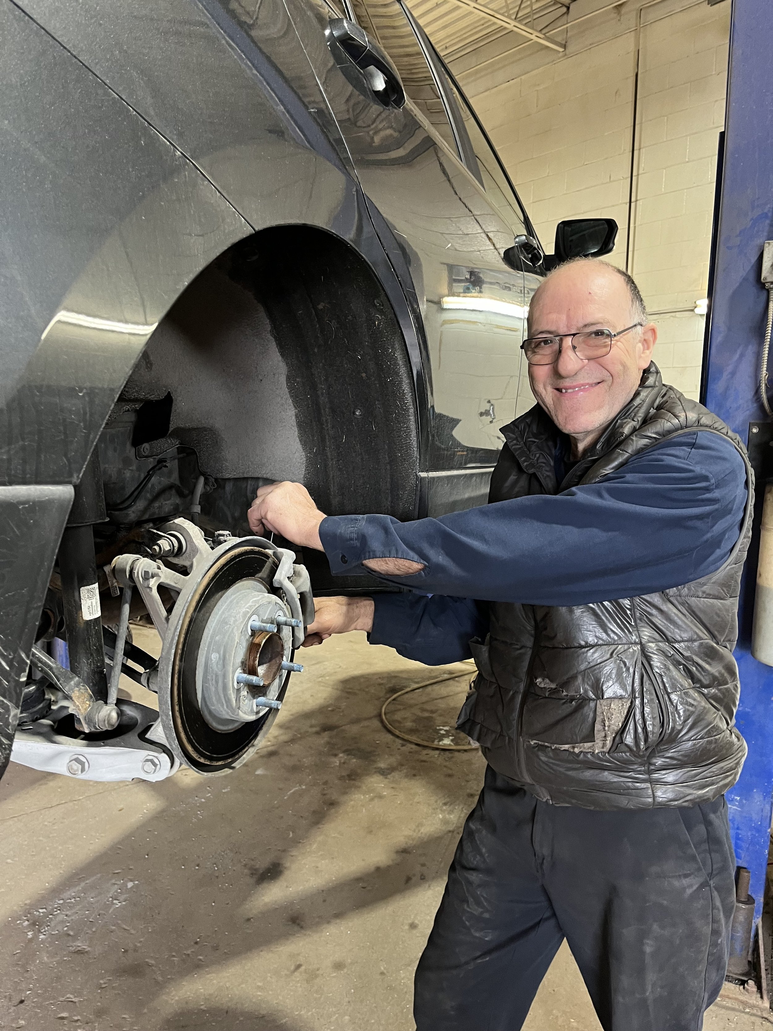 A man wearing glasses, a black vest over a blue shirt, and black pants is working on the front of a black car in a garage. The car's front wheel and brake system are exposed, and the man is holding a tool near the brake area. The garage has a concrete floor and yellow brick walls.