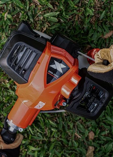 A person wearing gloves holding an orange and black power tool outdoors on green grass.