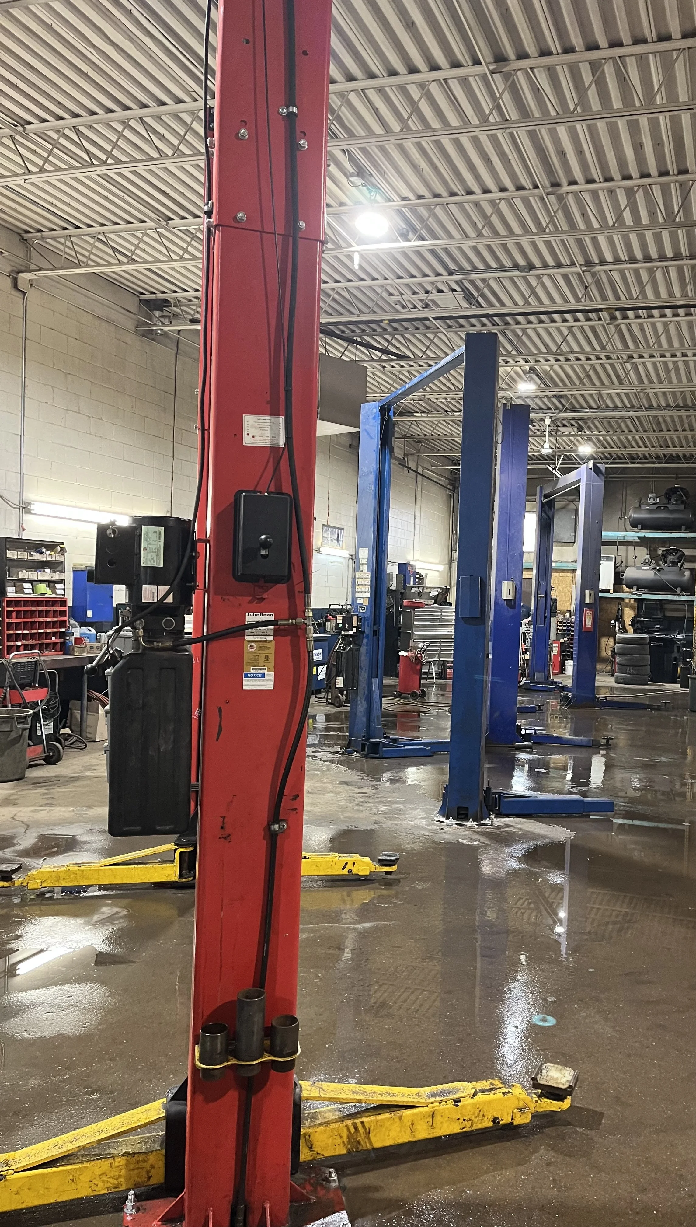 Inside an automotive repair shop with blue vehicle lifts, tools, and equipment, wet concrete floor, and overhead lighting.