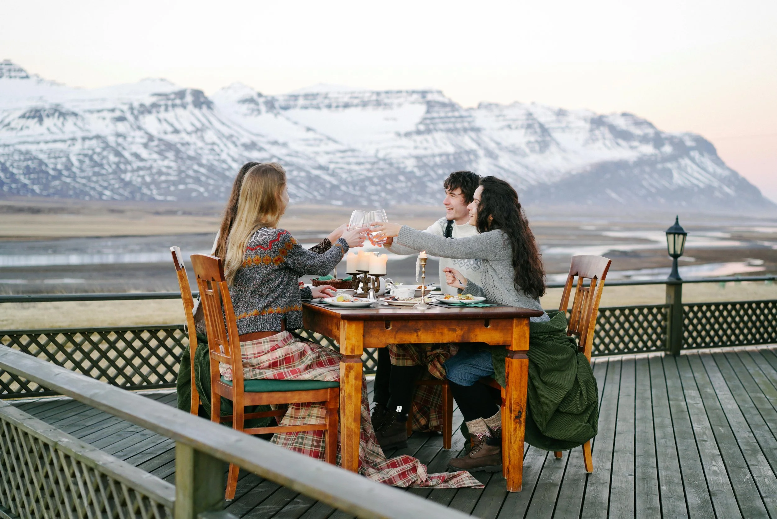 Four people sit at a wooden outdoor dining table, raising glasses in a toast, with snow-covered mountains in the background.