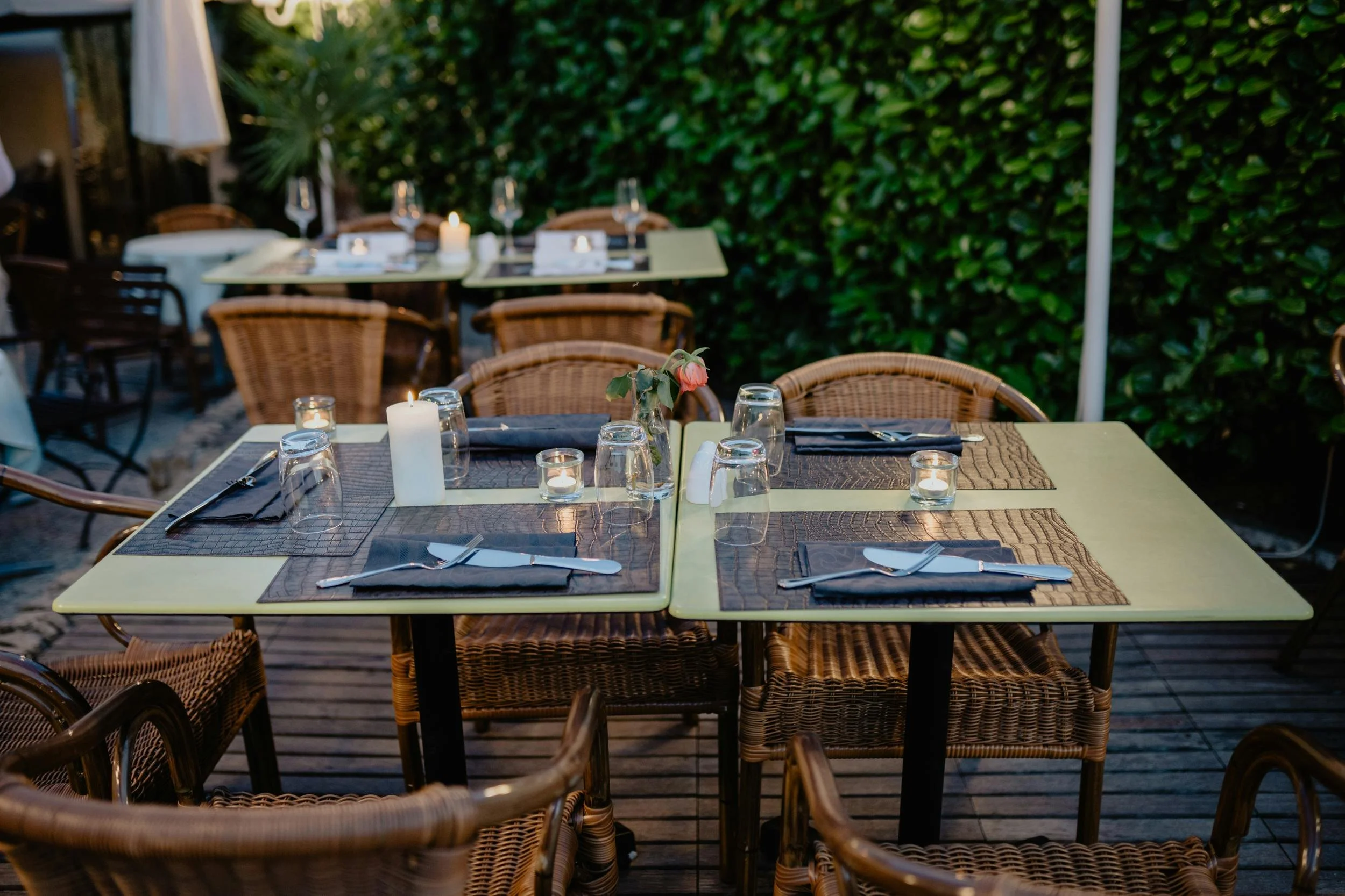 Outdoor dining tables set with candles, glasses, and napkins on a patio, surrounded by green hedge walls.