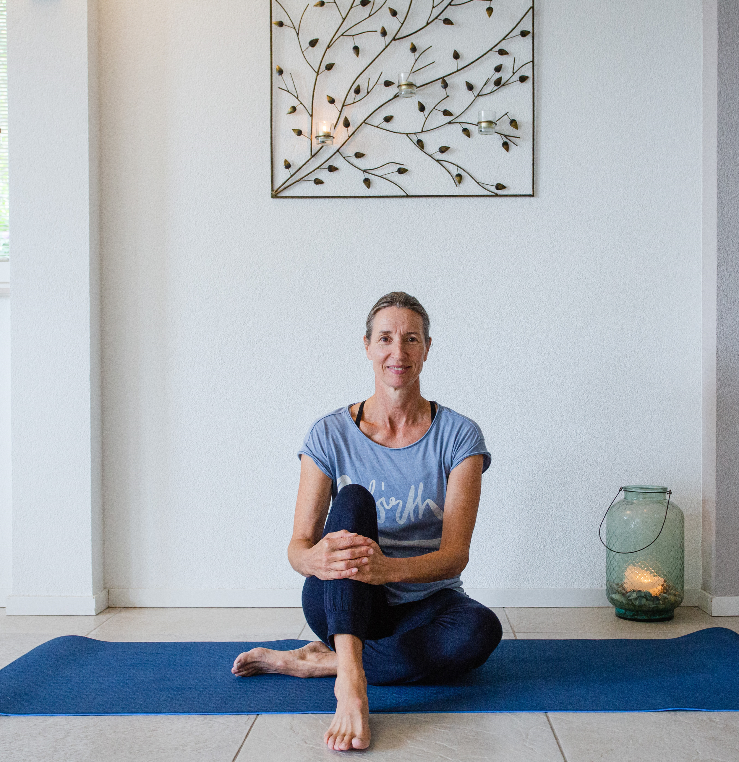Vrouw die een zittende yogapositie doet op een blauwe yogamat in een lichte kamer met wit metselwerk muur en een wanddecoratie met takken en bladeren