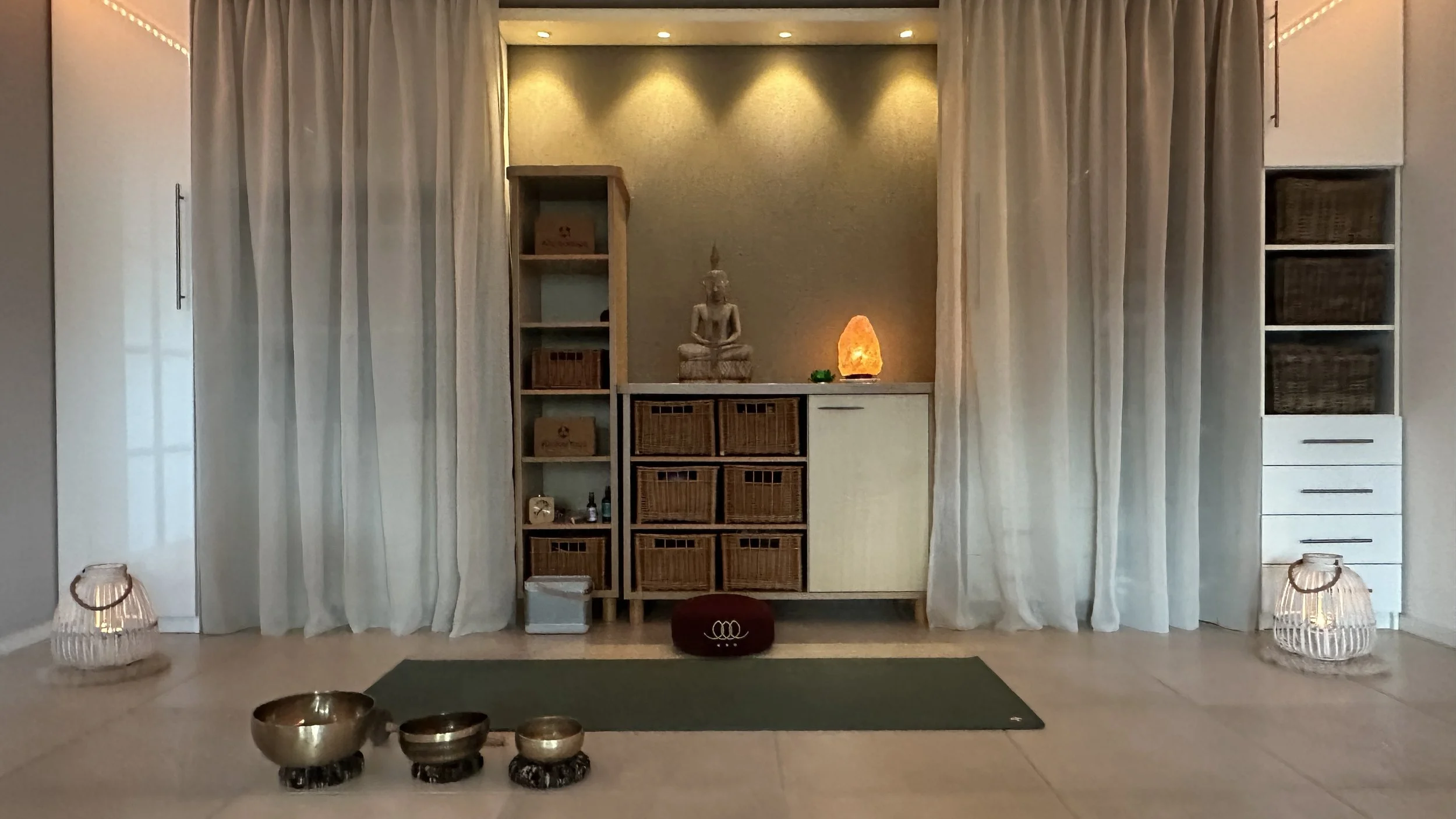 Een rustige kamer met een meditatiehoek, witte gordijnen, een houten boekenplank met manden, een beeld van een mediterende Boeddha, een himalaya zoutlamp, en traditionele tinkelende klankschalen op de vloer.