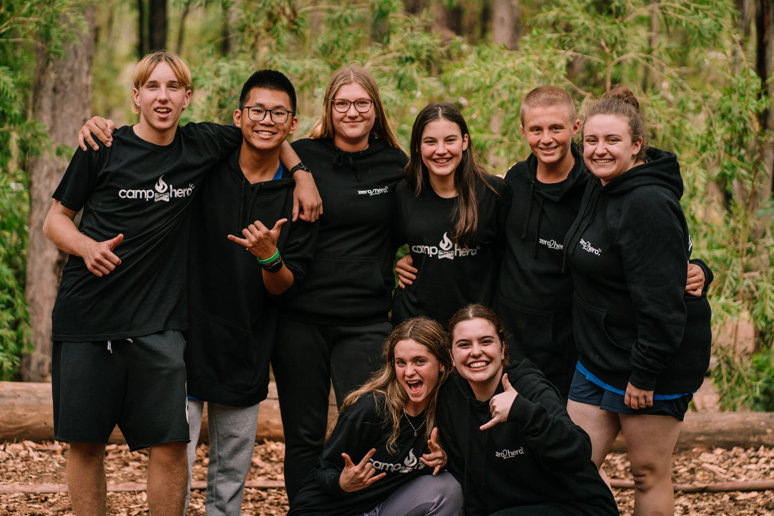 Group of smiling teenagers posing outdoors in a wooded area, some wearing black hoodies with camp logos.