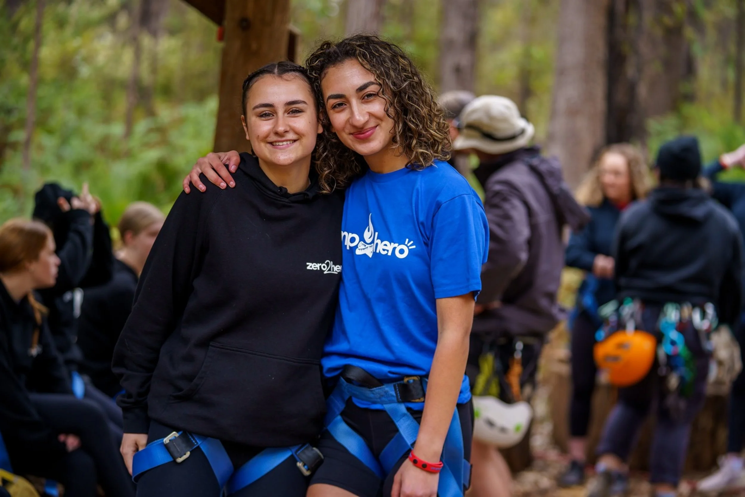 Two young women smiling and embracing outdoors in a wooded area, with other people in the background.