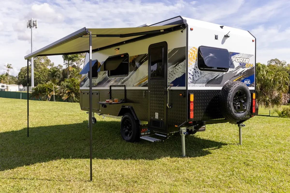 A modern RV with an extended awning, parked on a grassy field with palm trees in the background.
