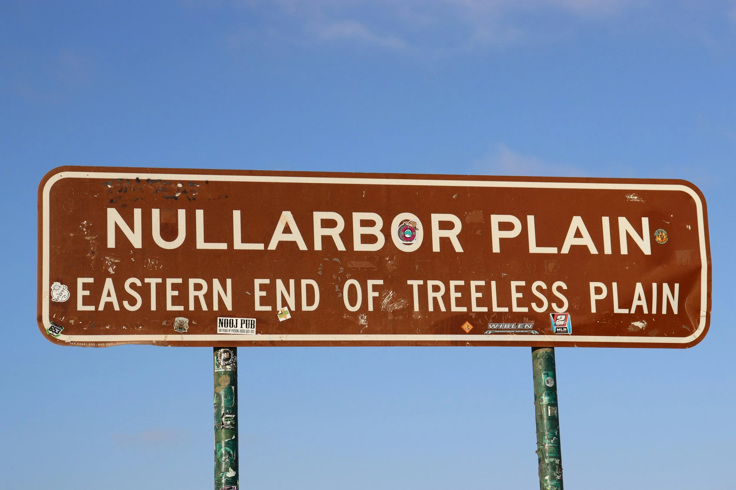 Brown street sign with white text reading "Nullarbor Plain" and additional information about the eastern end of treeless plain, covered in various stickers, against a blue sky.
