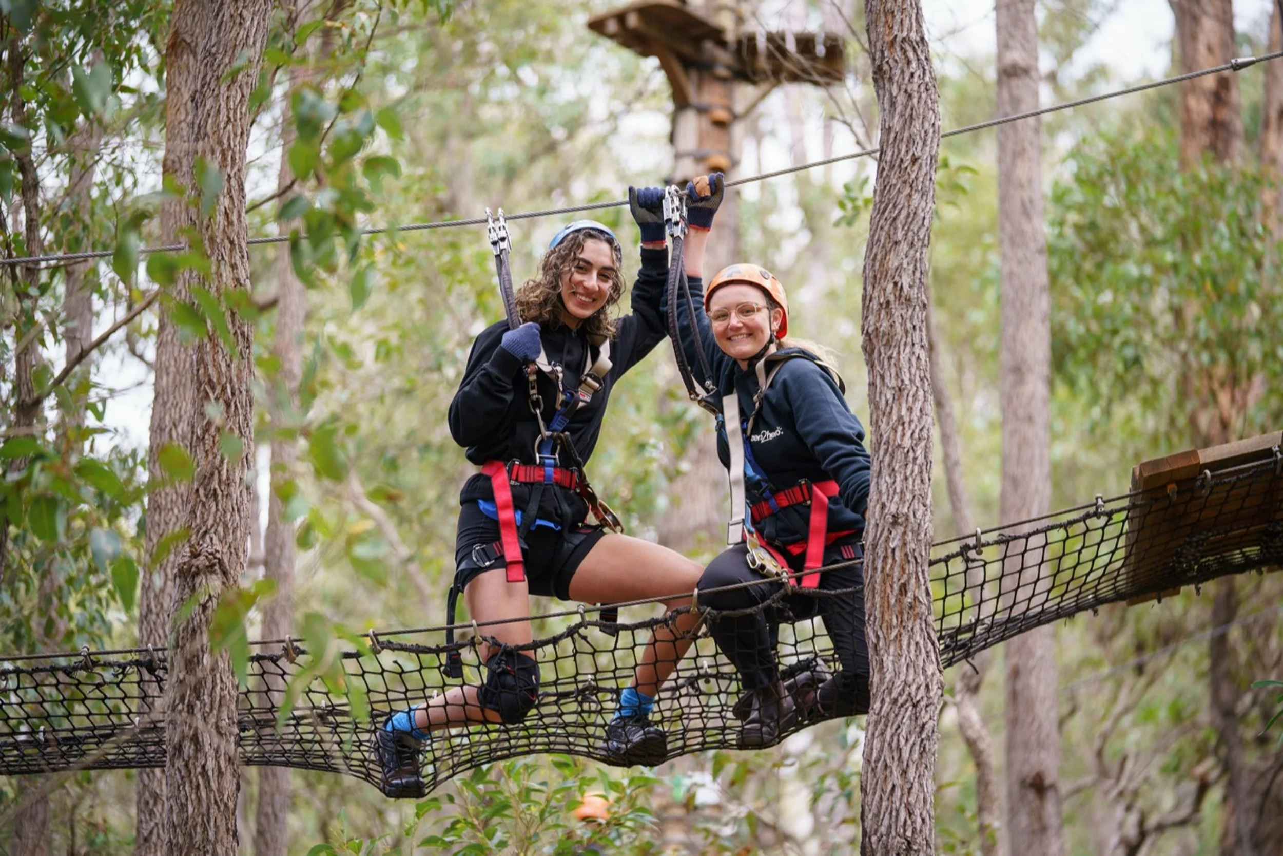 Two women on a suspension bridge in a forest, wearing helmets and harnesses, smiling at the camera.