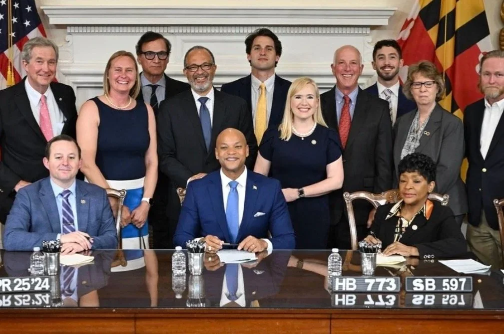 A group of diverse people, including men and women of different ethnicities and ages, gathered around a table with officials and documents. It appears to be a formal meeting or signing event, with some individuals smiling and looking at the camera. There are flags and a fireplace mantel in the background.