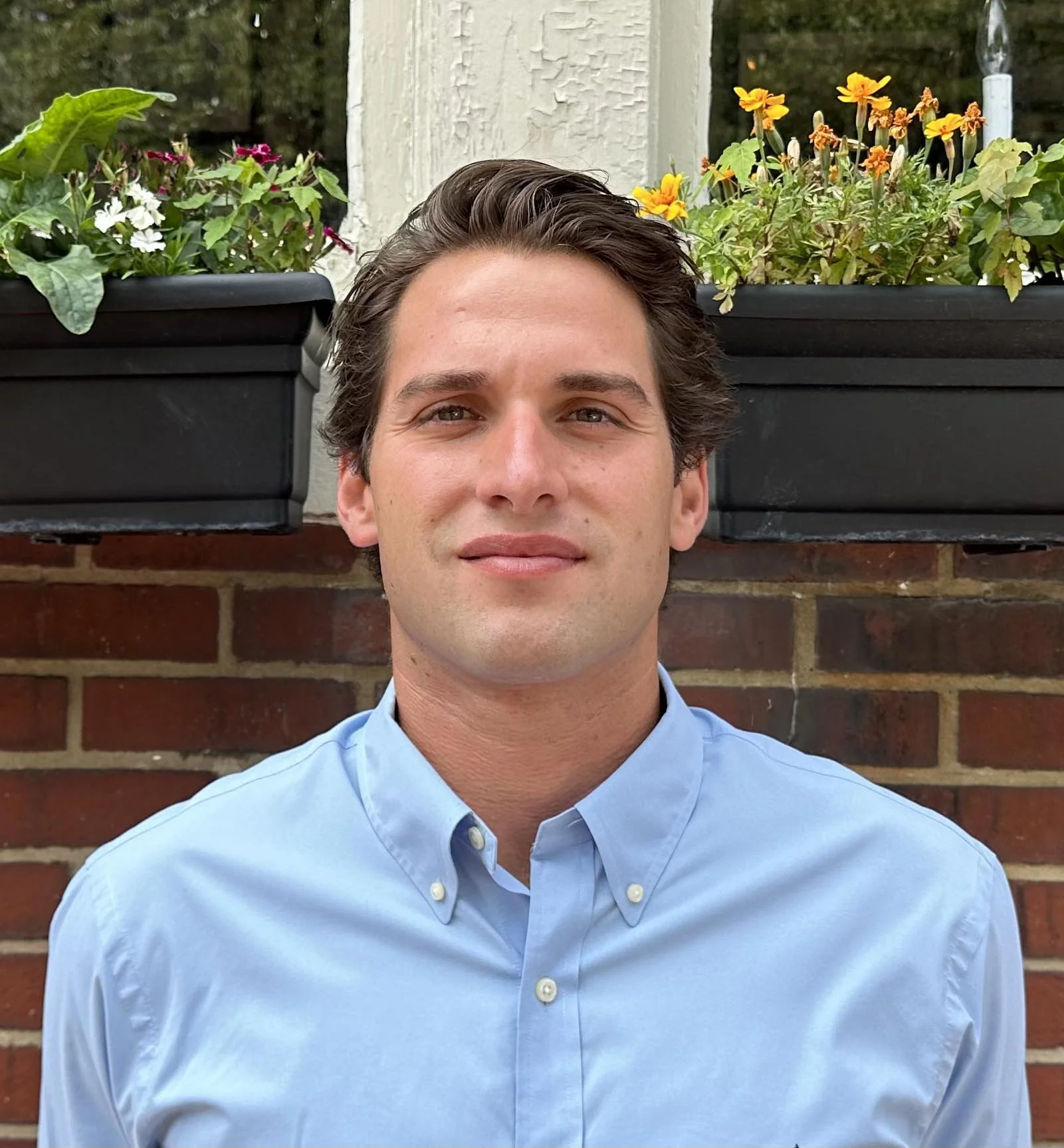A young man with dark hair wearing a light blue button-up shirt standing outside in front of a brick wall with two flower boxes filled with colorful flowers and green plants on a porch or balcony.