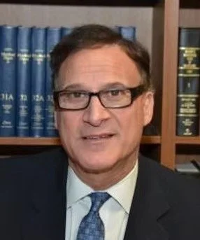 A man with glasses and dark hair, wearing a suit and tie, standing in front of a bookshelf filled with blue and dark-colored books.