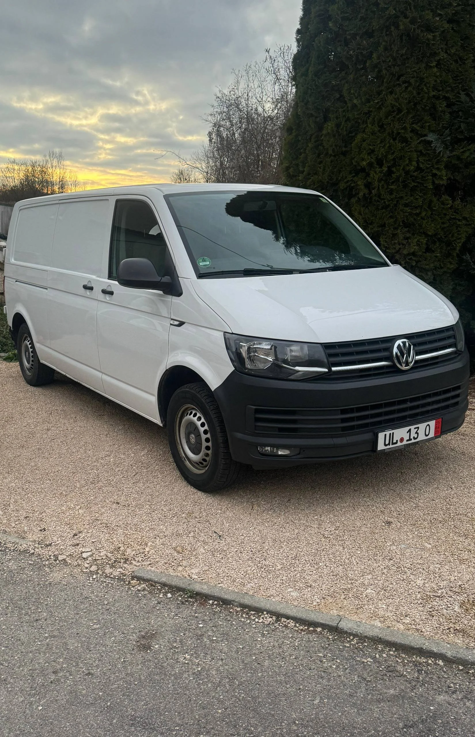 Ein weißer VW Transporter Park auf einer gepflasterten Fläche, im Hintergrund Bäume und ein bewölkter Himmel bei Sonnenuntergang.
