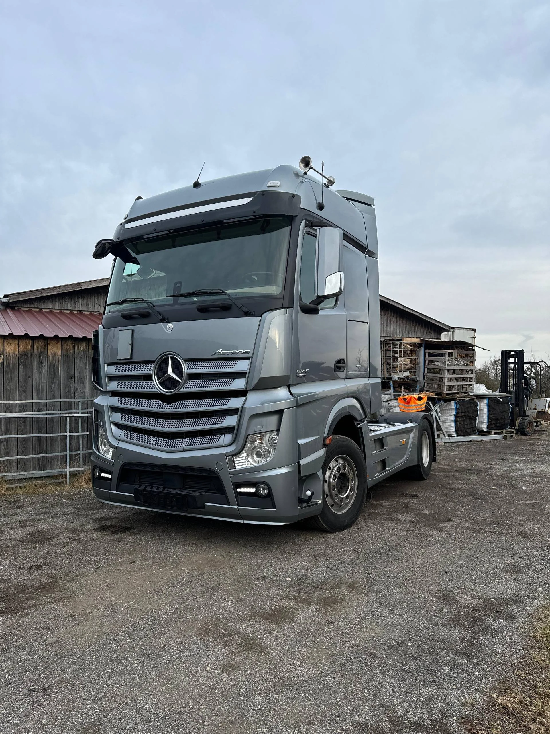 Ein grauer Mercedes-Benz LKW steht auf einem unbefestigten Parkplatz mit erkennbaren Holzstrukturen und einem Gabelstapler im Hintergrund. Der Himmel ist bewölkt.
