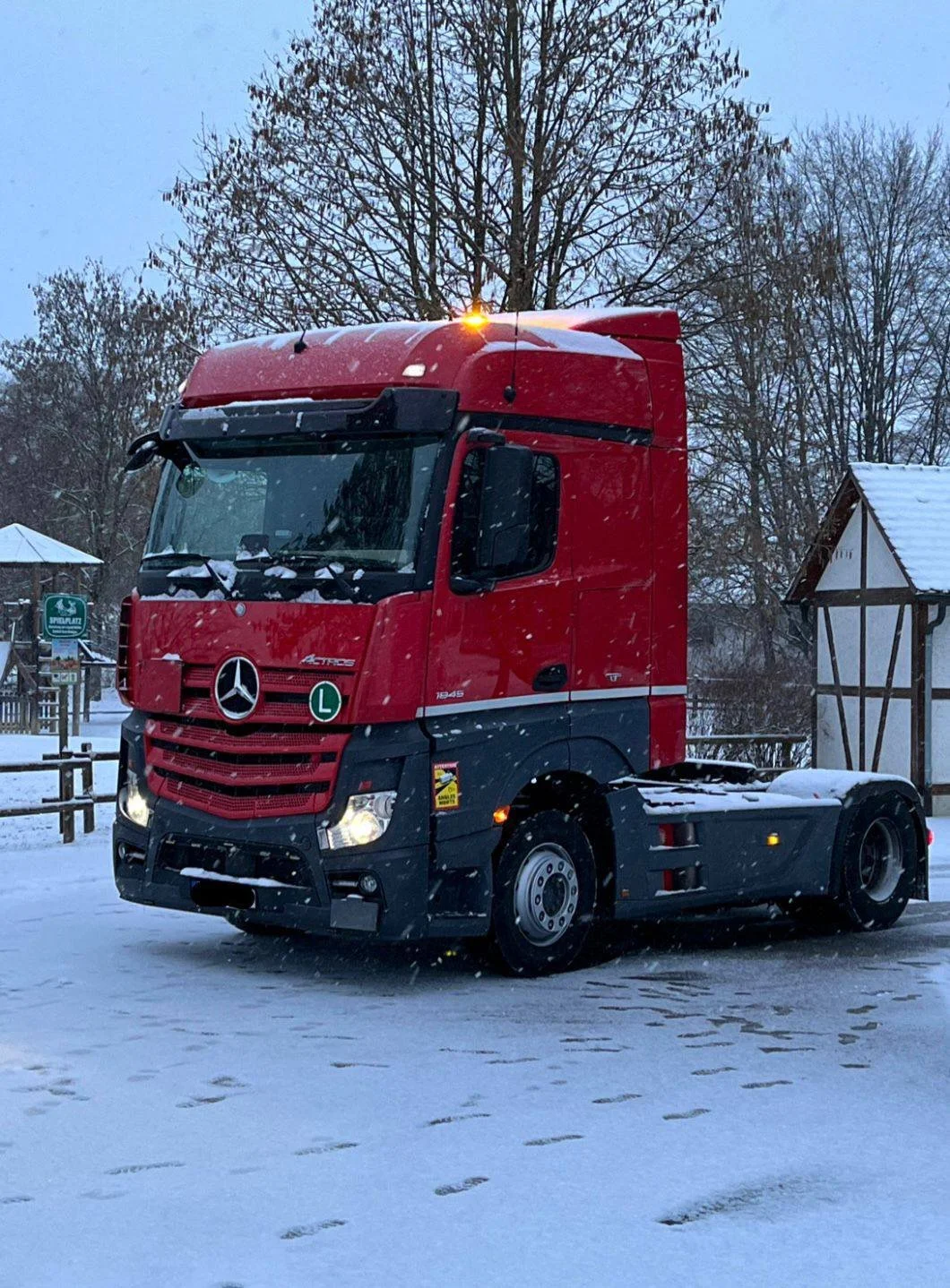 Ein roter Mercedes-Benz LKW auf verschneitem Boden, umgeben von Bäumen ohne Blätter in winterlicher Landschaft.