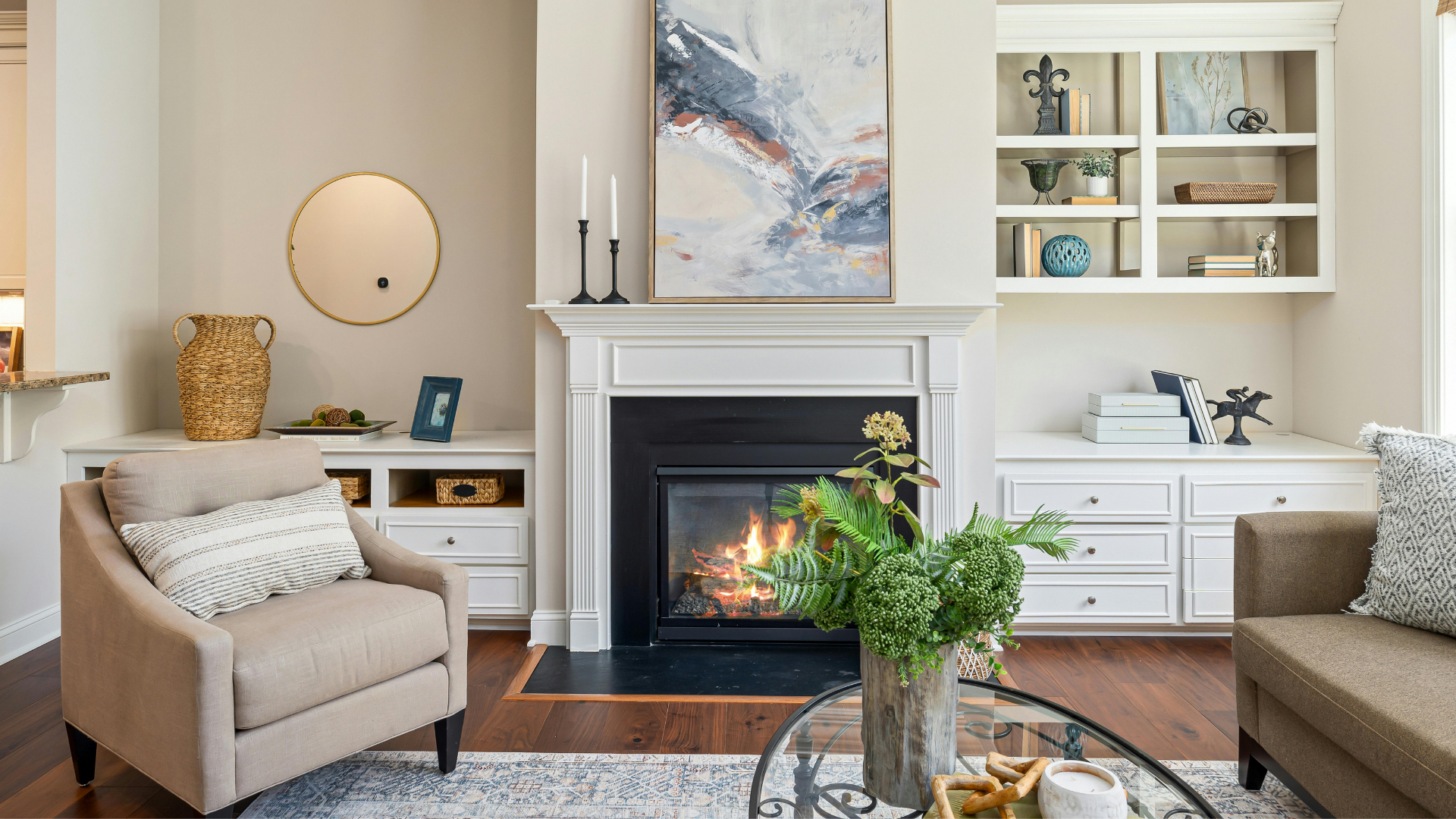 A cozy living room with a fireplace, beige armchair with a striped pillow, glass-top coffee table with decorative objects, built-in white shelves with books and decor, abstract painting above fireplace, and hardwood flooring.