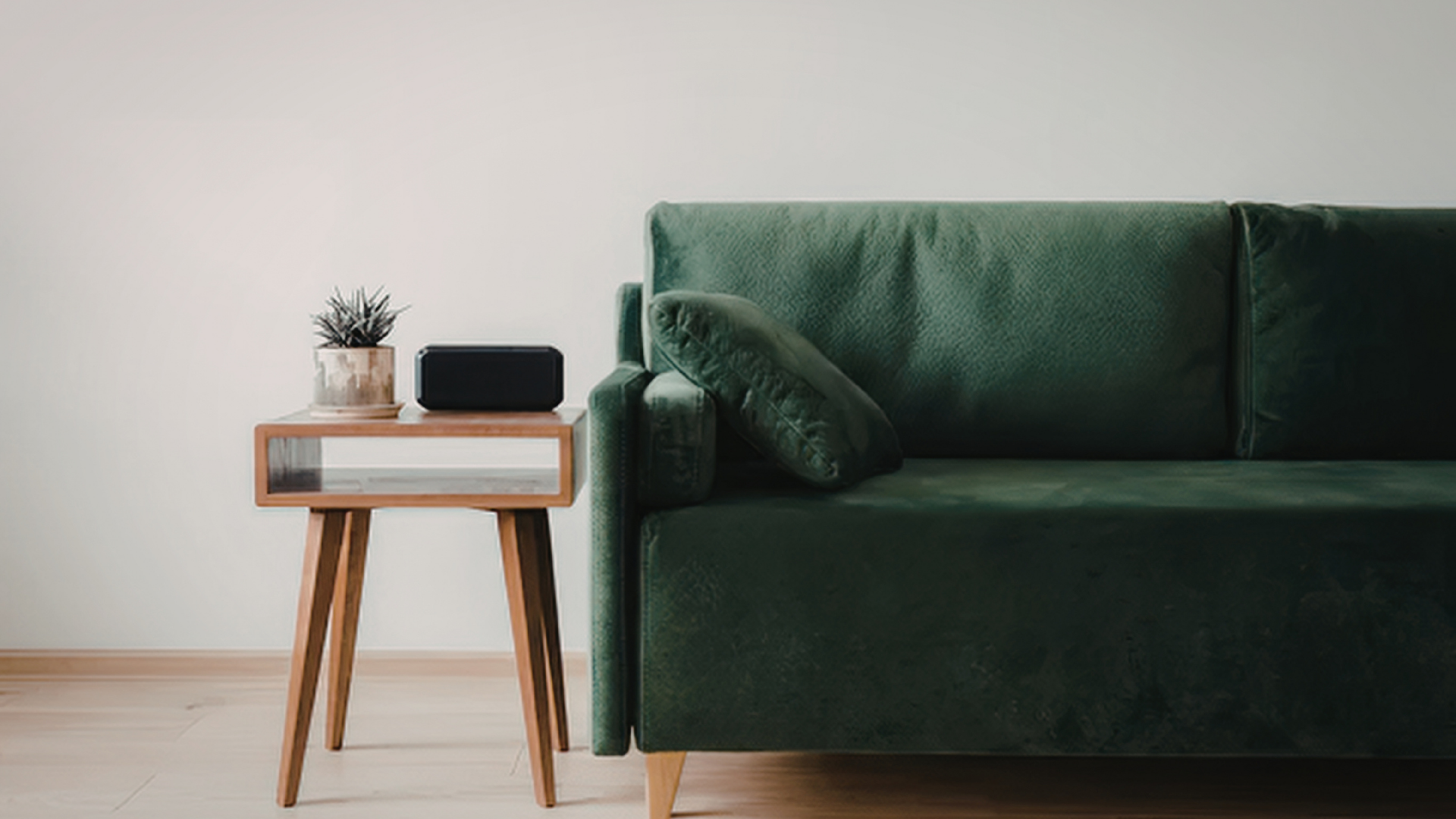 Living room with a green velvet sofa and a wooden side table with a potted plant and a black speaker on it.