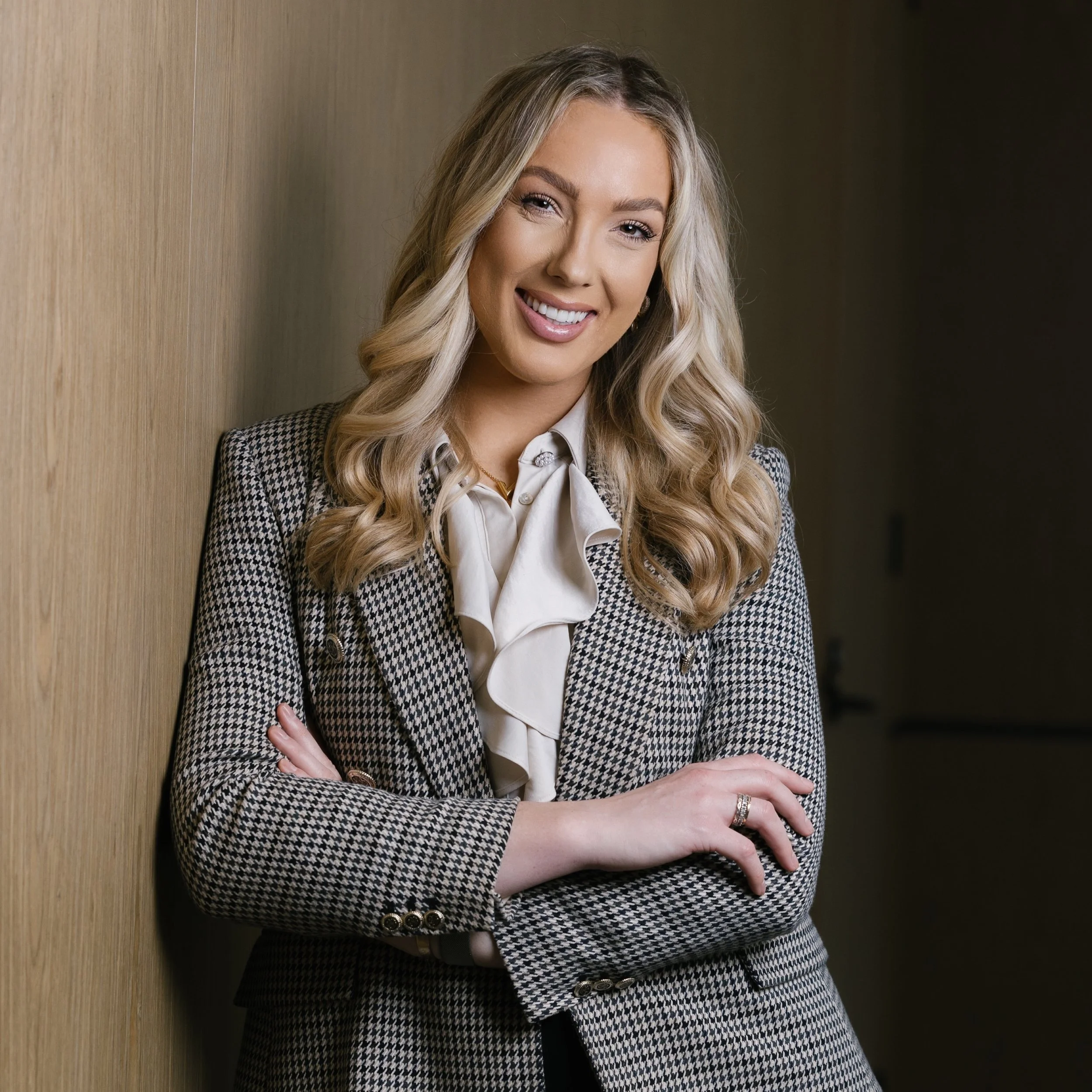 A woman with long blonde hair styled in loose waves, smiling and crossing her arms, wearing a houndstooth blazer over a cream blouse with a bow tie, standing against a wooden wall.