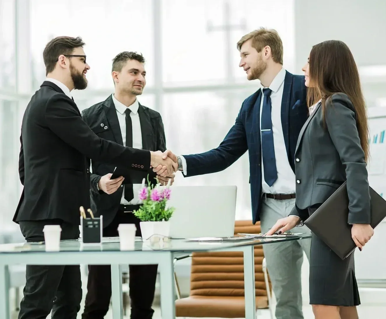 Five business professionals, three men and two women, in formal business attire, shaking hands and engaging in conversation in a modern, well-lit office space. One woman is holding a folder, and the table in front of them has a flowerpot, glasses, papers, and office supplies.